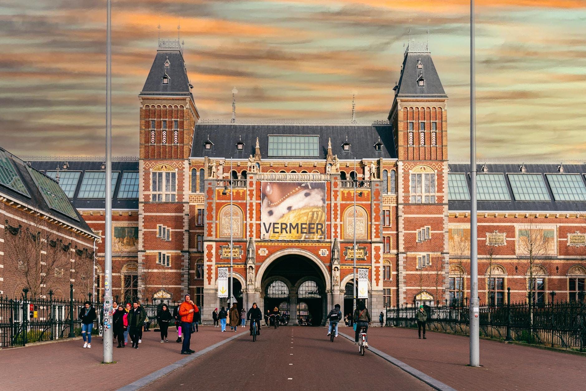 Vista frontal del Rijksmuseum en Ámsterdam con personas caminando y en bicicleta bajo un cielo nublado de invierno. Pancarta de Vermeer en exhibición.