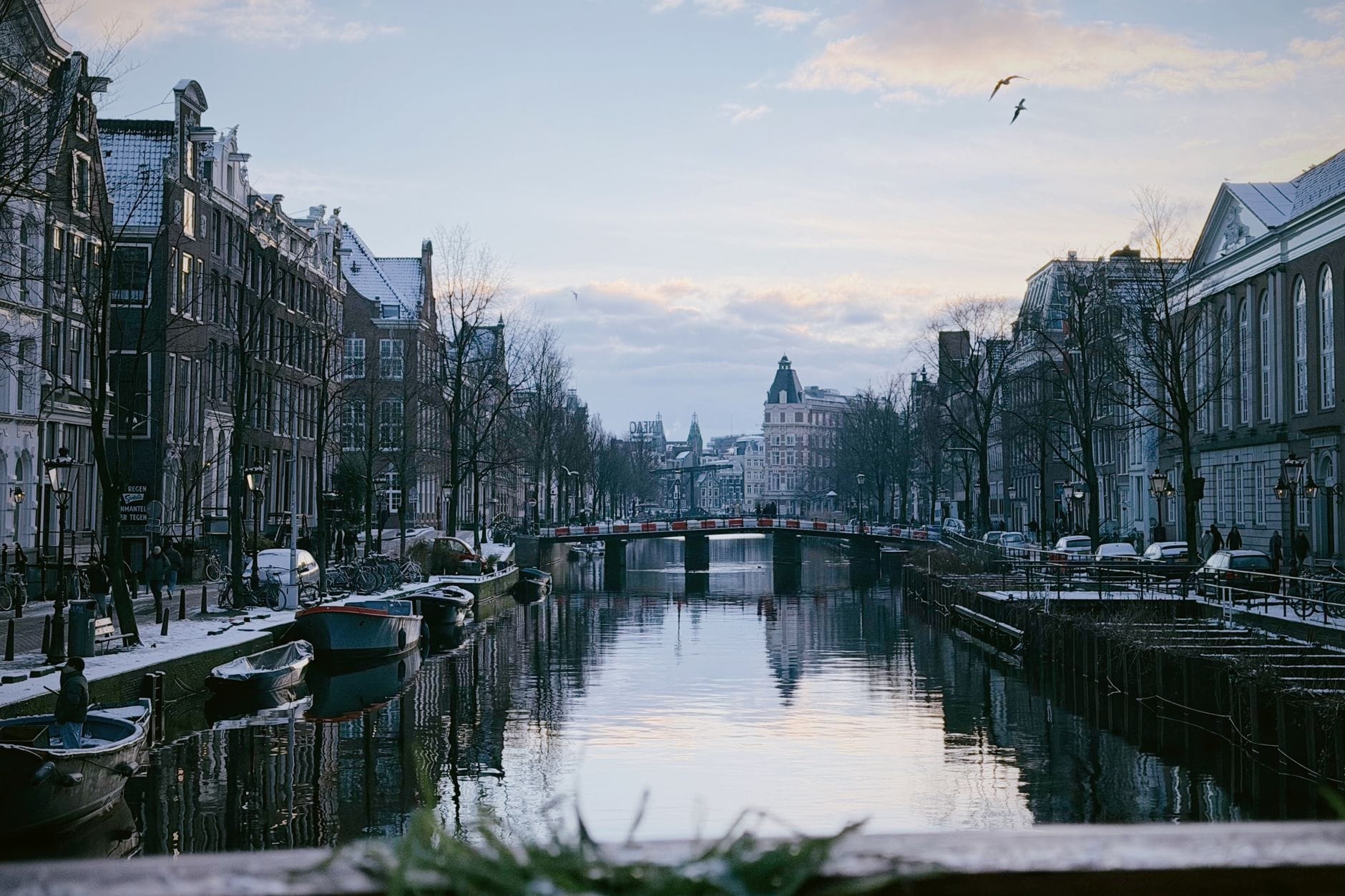 Escena invernal en Ámsterdam con árboles sin hojas, casas históricas junto al canal, barcas y un puente al anochecer, con algo de nieve visible en los tejados.