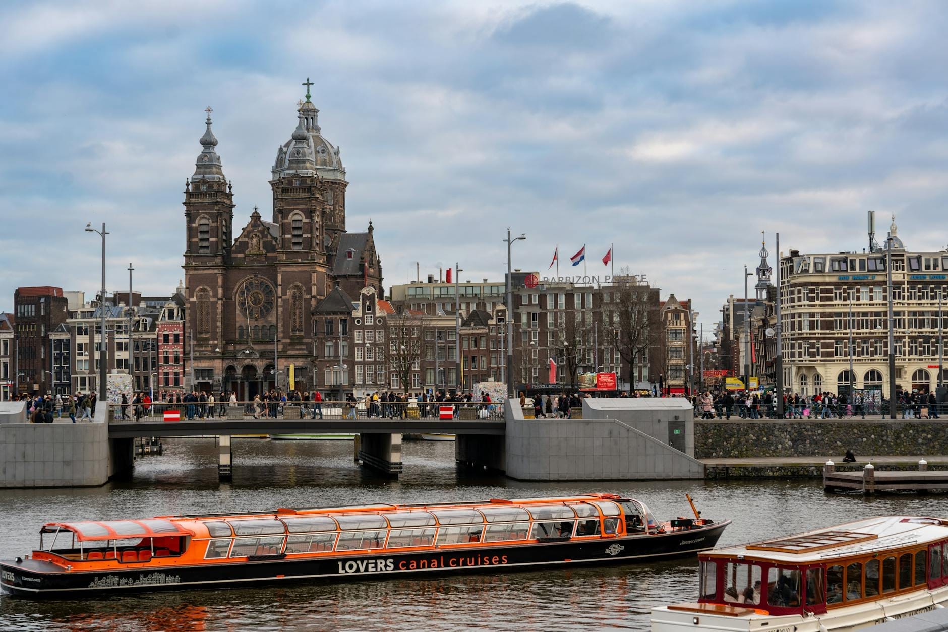 Barcos de crucero por el canal en el centro de Ámsterdam con la histórica Basílica de San Nicolás y antiguos edificios de la ciudad al fondo.