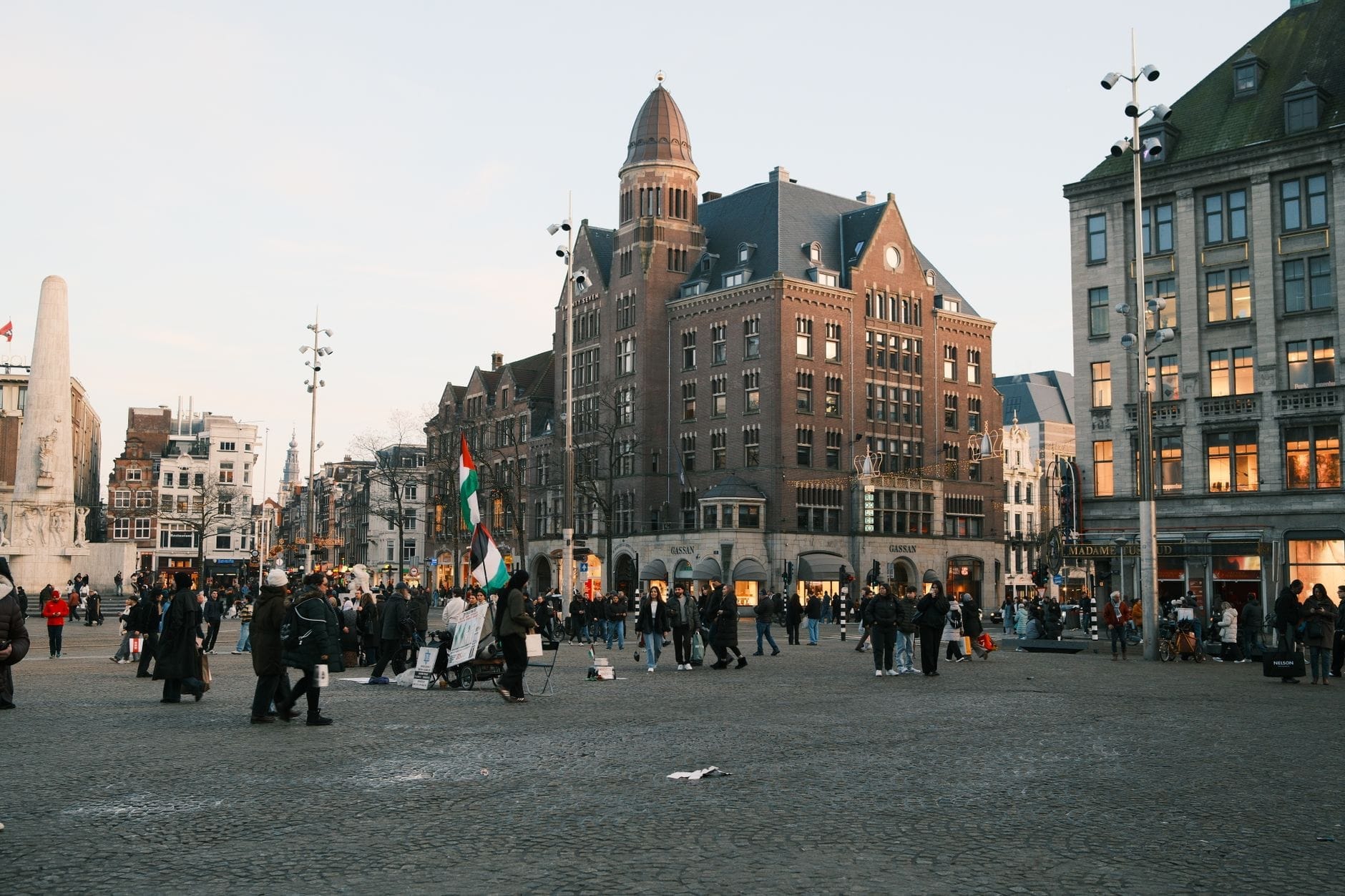 Plaza Dam en Ámsterdam con personas caminando y edificios históricos visibles, incluida la reconocible torre puntiaguda del NH Grand Hotel Krasnapolsky.