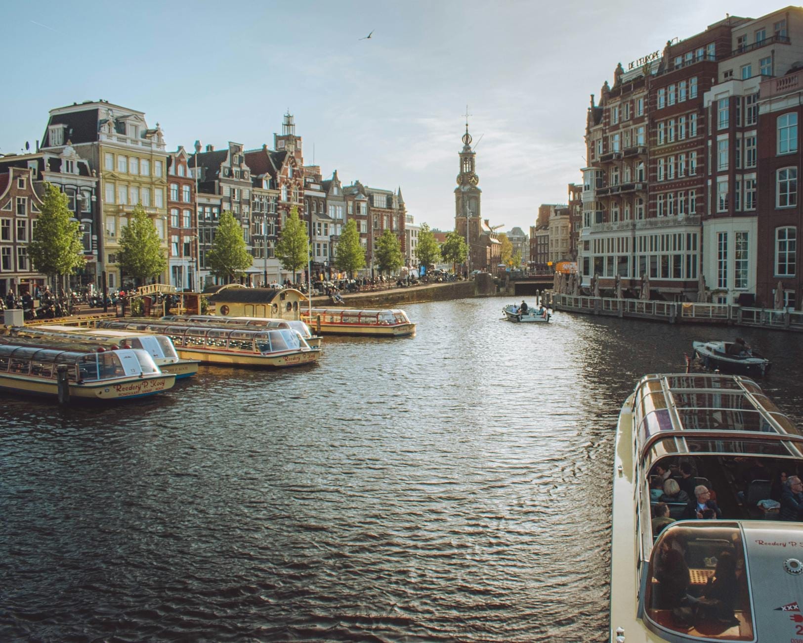 Amplia escena de un canal en Ámsterdam con sus icónicas casas estrechas, barcas y una histórica torre del reloj bajo un cielo azul. Una imagen llena de vida y ambiente urbano.