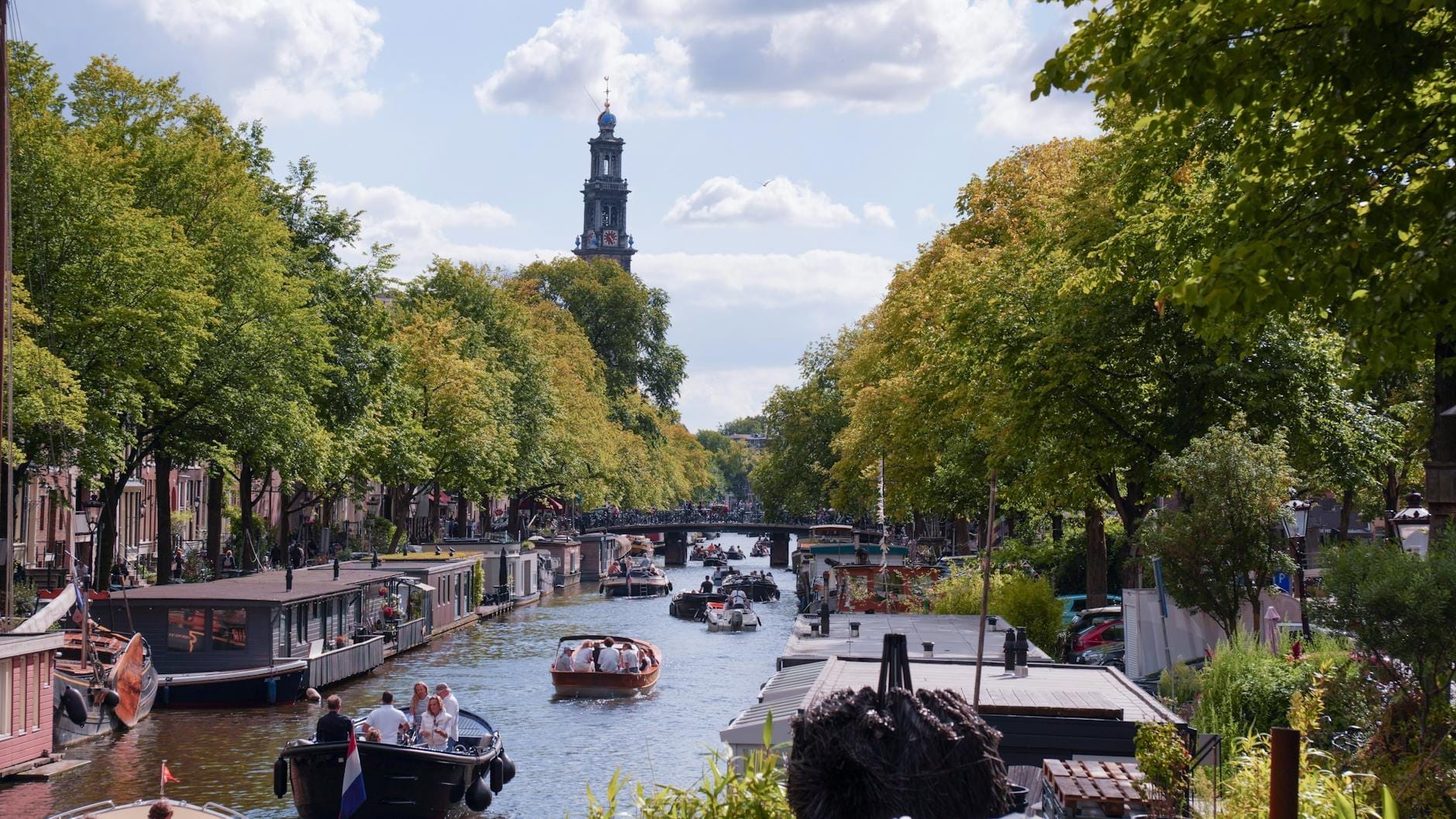 Vista del canal de Ámsterdam con varios barcos, frondosos árboles, casas flotantes y la torre de la Westerkerk al fondo bajo un cielo azul.