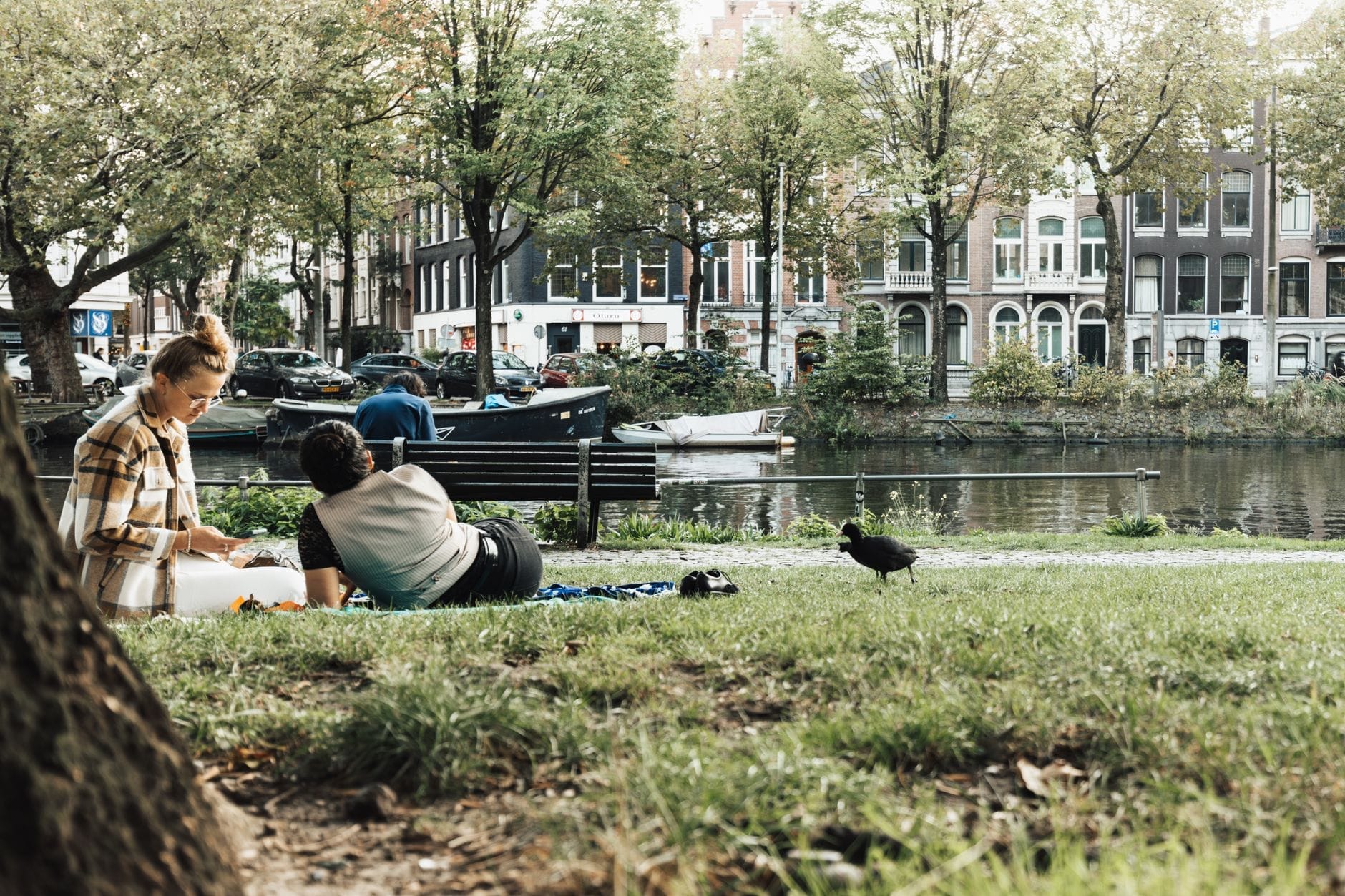 Dos personas relajándose y haciendo un picnic en el césped junto a un canal de Ámsterdam, con casas del canal y botes al fondo.