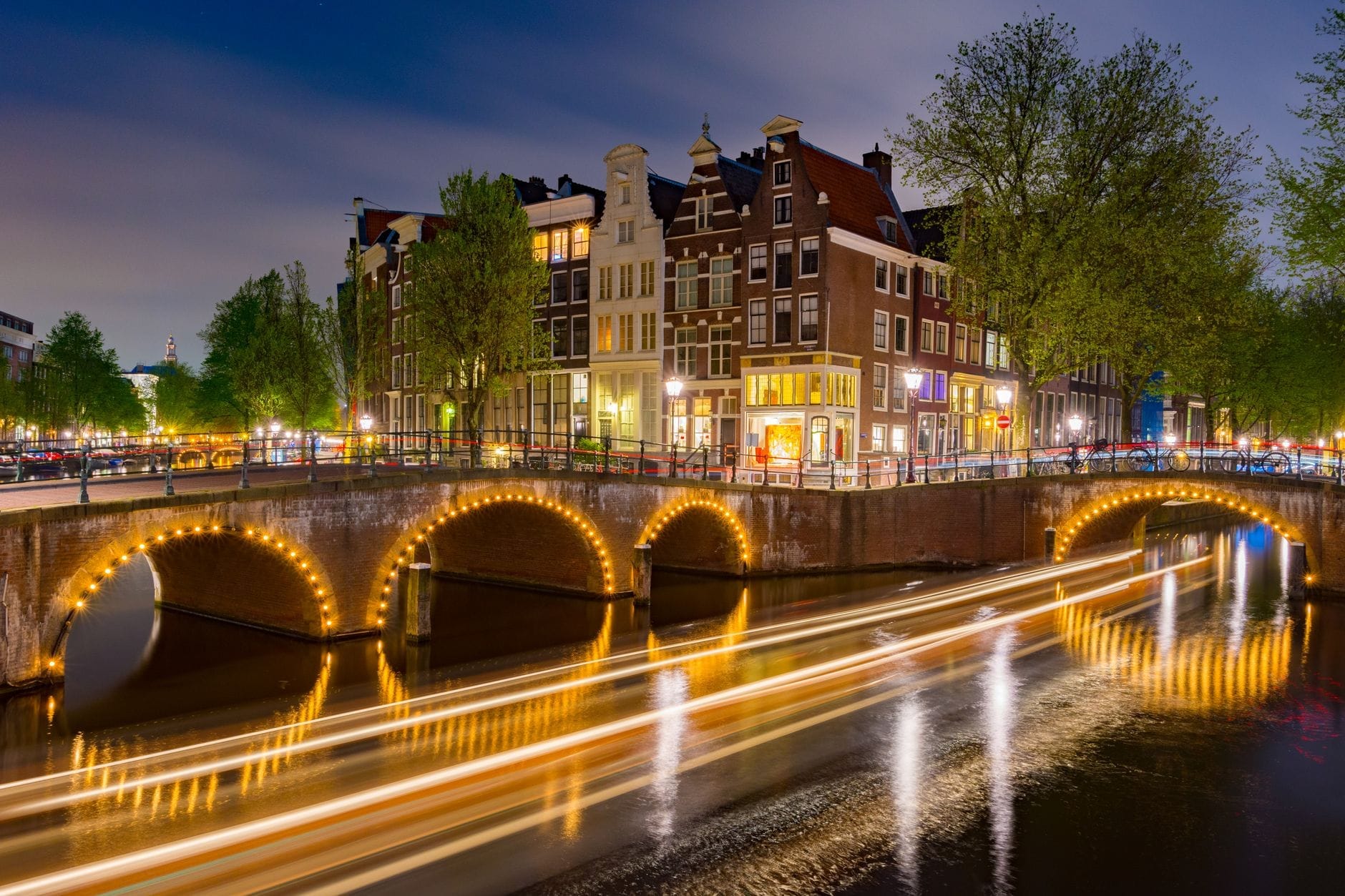 Canal de Ámsterdam de noche con casas históricas, puentes iluminados y estelas de luz de un barco en movimiento, capturando un ambiente festivo nocturno.