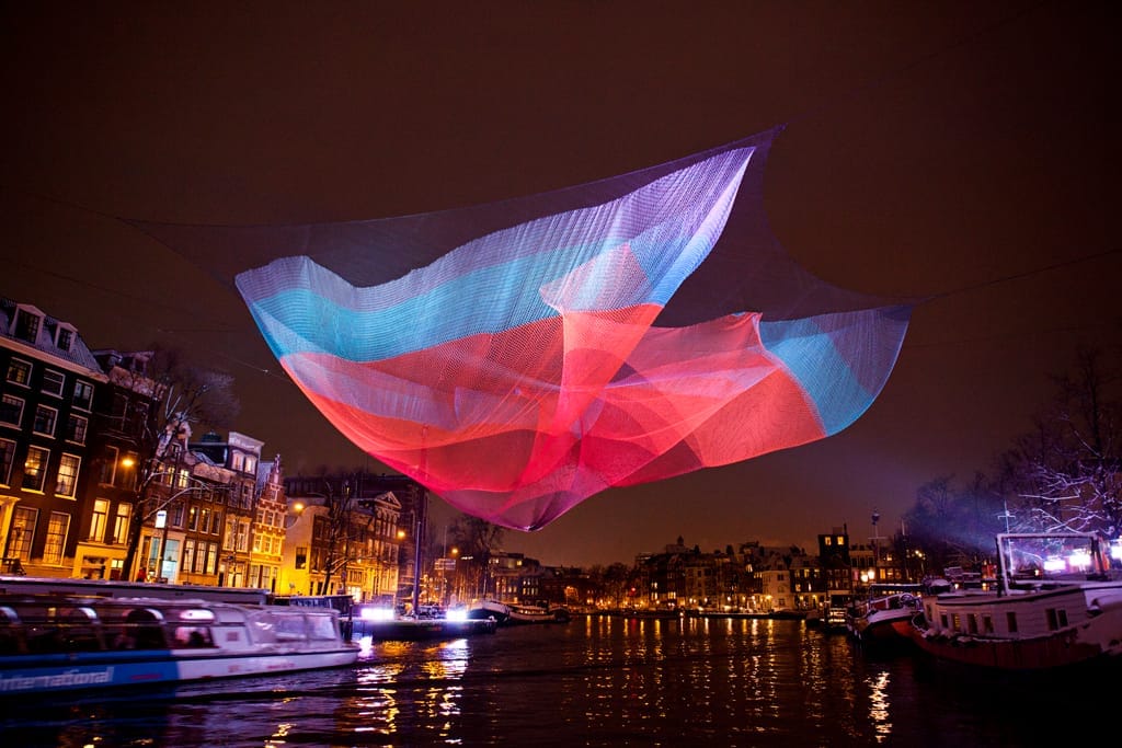 Colorida instalación de arte de red iluminada sobre un canal de Ámsterdam por la noche, con edificios históricos y barcos turísticos visibles durante el Light Festival.