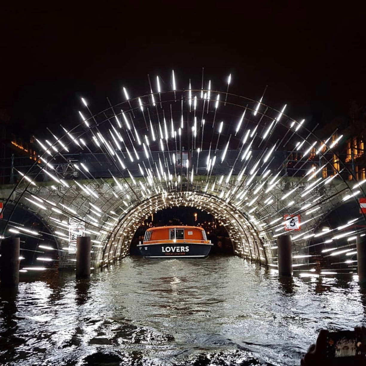 Instalación de arte iluminada en el Amsterdam Light Festival, con un barco pasando bajo un puente iluminado con brillantes luces blancas sobre el canal por la noche.