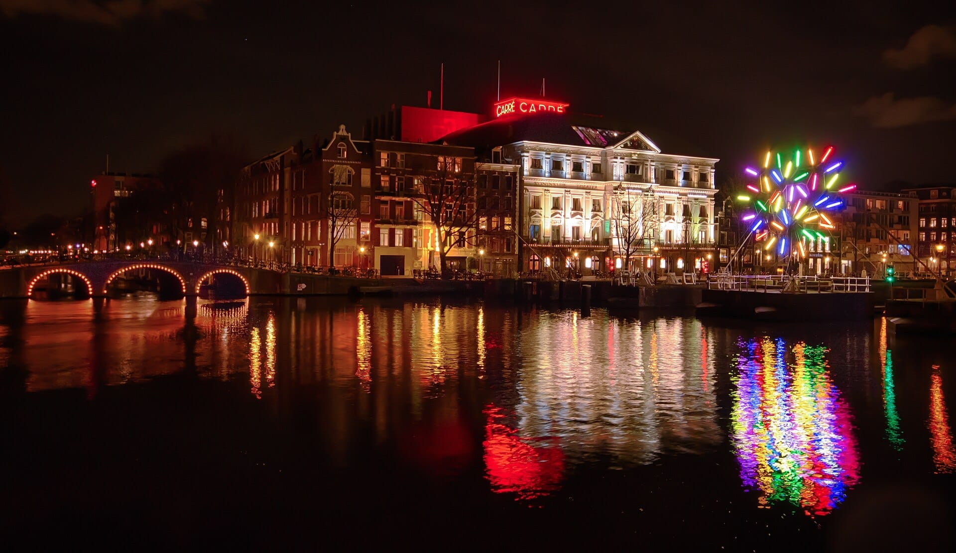 Edificios históricos iluminados y puente sobre el canal reflejados en el agua durante el Amsterdam Light Festival, con una colorida instalación de luces en la orilla.