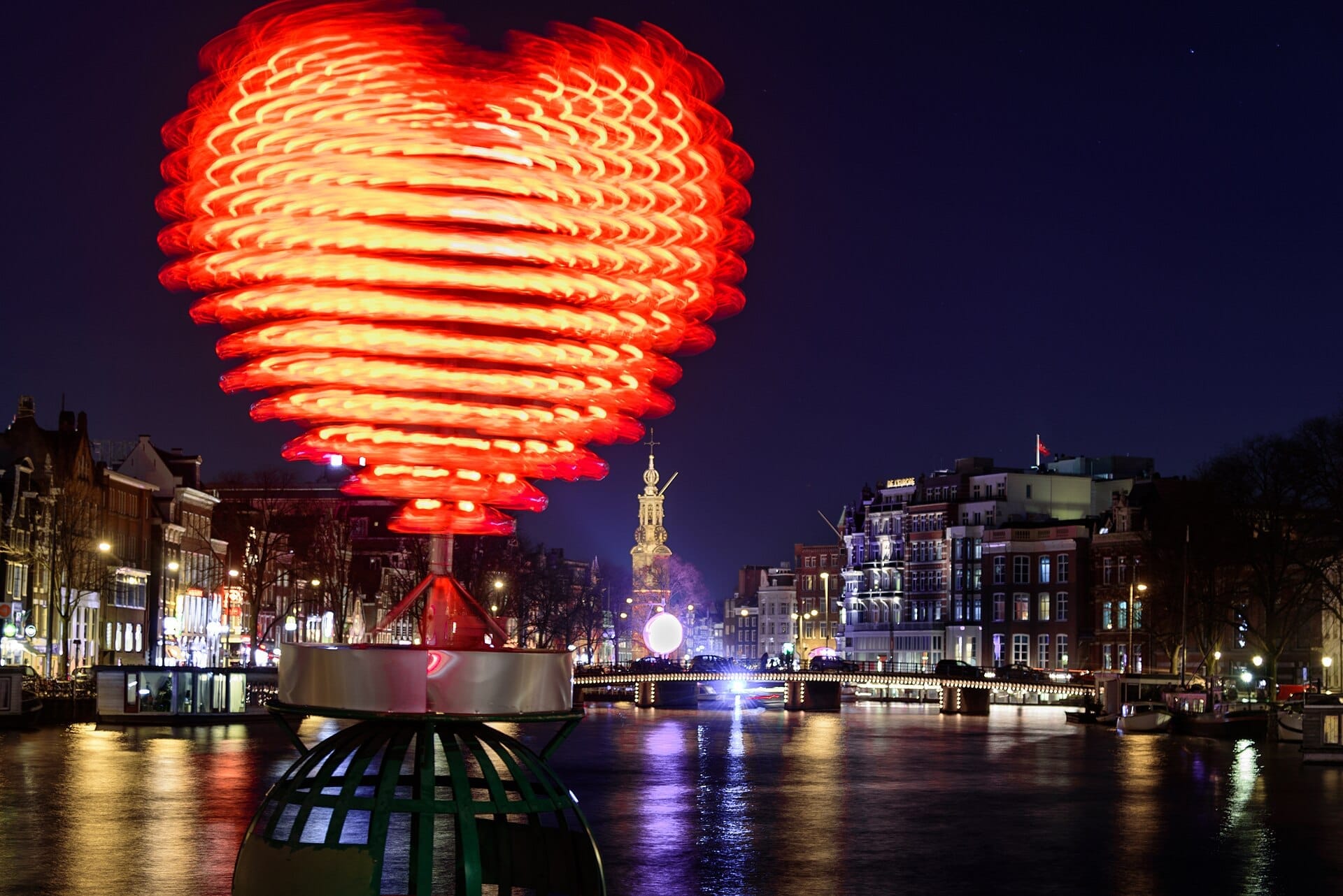 Una gran escultura luminosa en forma de corazón iluminada brilla sobre un canal de Ámsterdam de noche durante el Amsterdam Light Festival, con edificios históricos y reflejos en el agua.