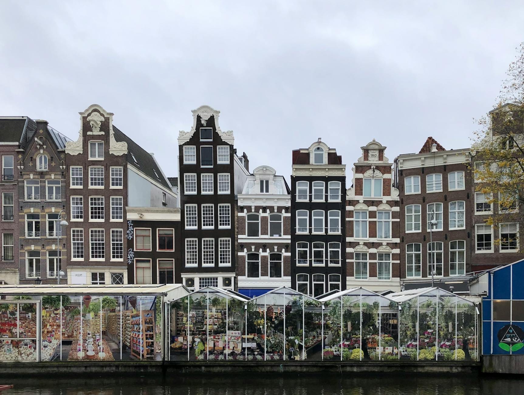 Cảnh những ngôi nhà ven kênh đặc trưng của Amsterdam phía sau các quầy hoa nổi nổi tiếng của Bloemenmarkt trên kênh Singel, dưới bầu trời nhiều mây.