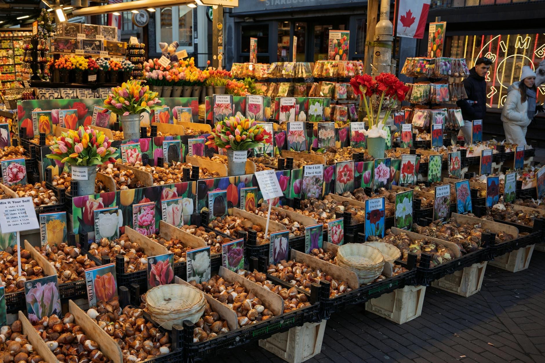 Một quầy chợ sôi động ở Amsterdam bán củ hoa và bó hoa, với những gói hàng đầy màu sắc được trưng bày và người mua sắm đang dạo xem phía sau.
