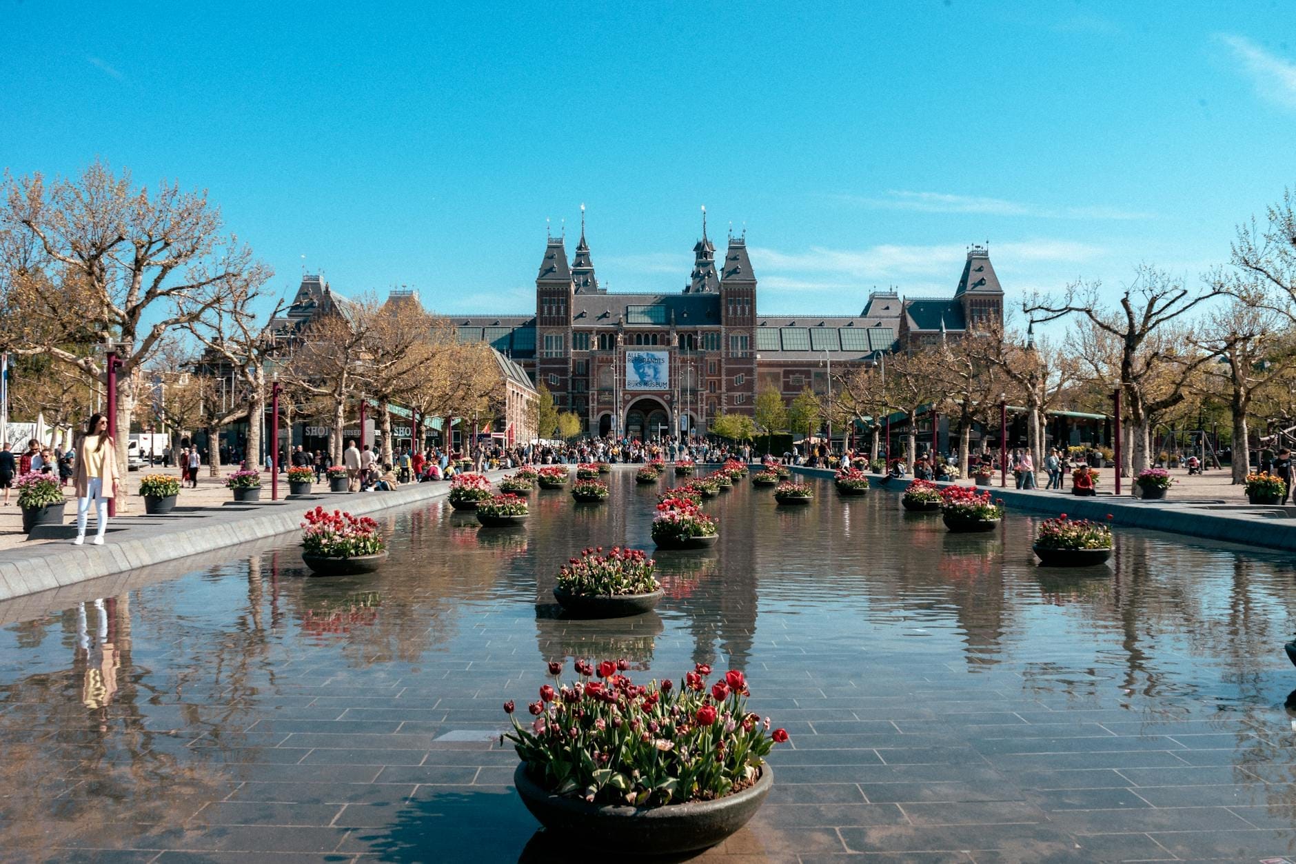 Góc nhìn rộng của Rijksmuseum ở Amsterdam với các chậu hoa tulip trong hồ phản chiếu và người dân tụ tập xung quanh, làm nổi bật khung cảnh địa danh văn hóa sôi động.