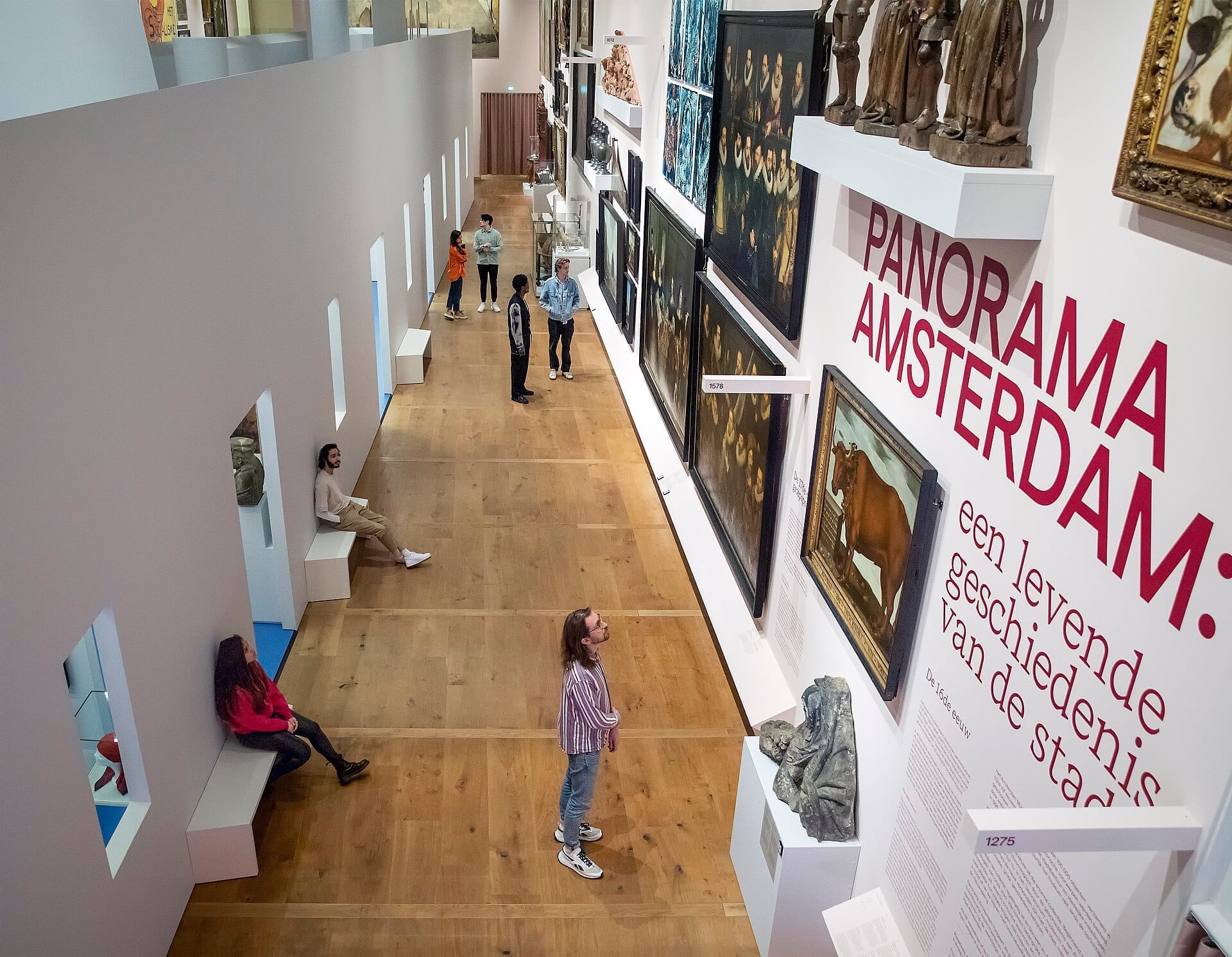 Los visitantes exploran la galería del Amsterdam Museum, admirando pinturas históricas y artefactos expuestos en paredes blancas con suelos de madera y bancos modernos.
