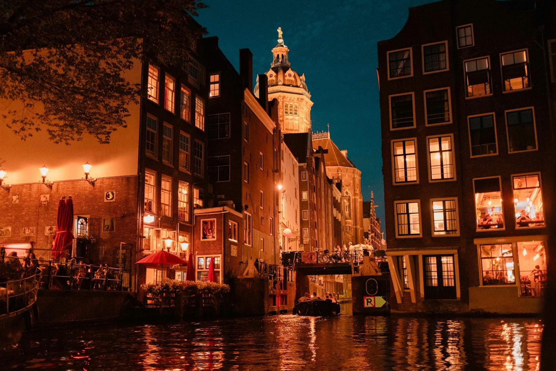 Canal de Amsterdam de noche con bares y restaurantes iluminados, personas sentadas al aire libre y una torre de iglesia al fondo.