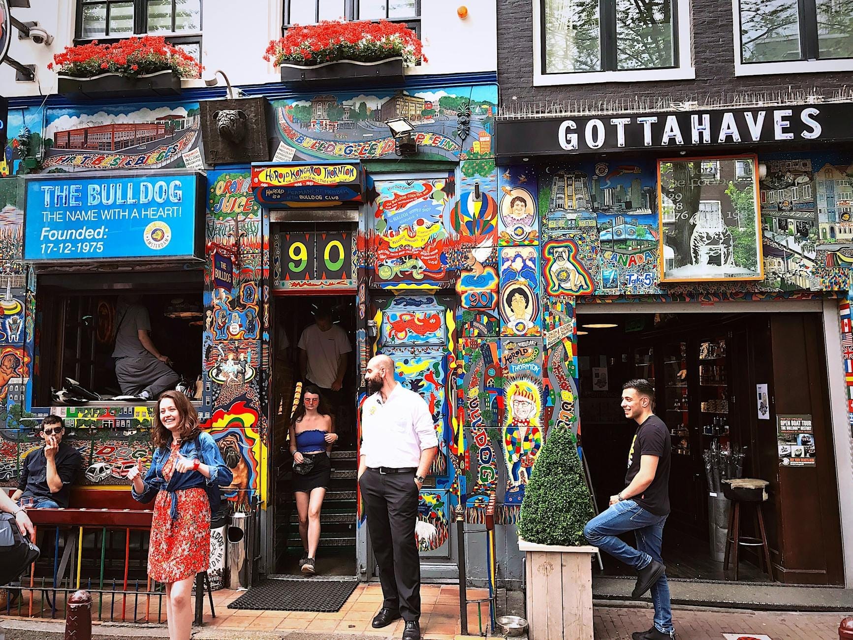 Exterior colorido del coffeeshop The Bulldog en Ámsterdam con personas frente a la entrada y vibrantes murales que cubren la fachada.