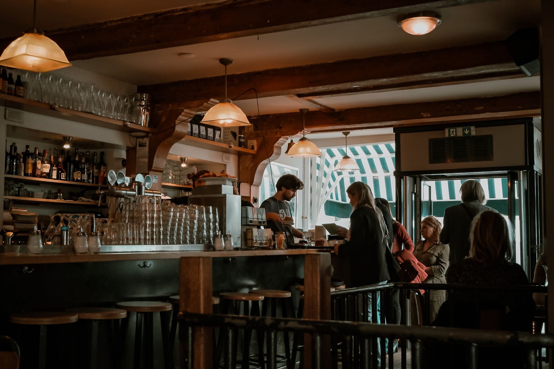 Interior de bar cálidamente iluminado con vigas de madera, taburetes, cristalería y un bartender sirviendo a clientes, creando un ambiente clásico de café marrón.