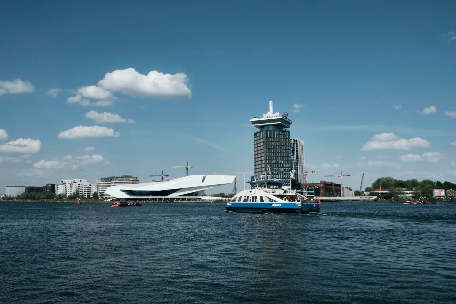 Ferri cruzando el río IJ con el EYE Filmmuseum y la Torre A'DAM visibles en el paseo marítimo de Amsterdam-Noord bajo un cielo azul.