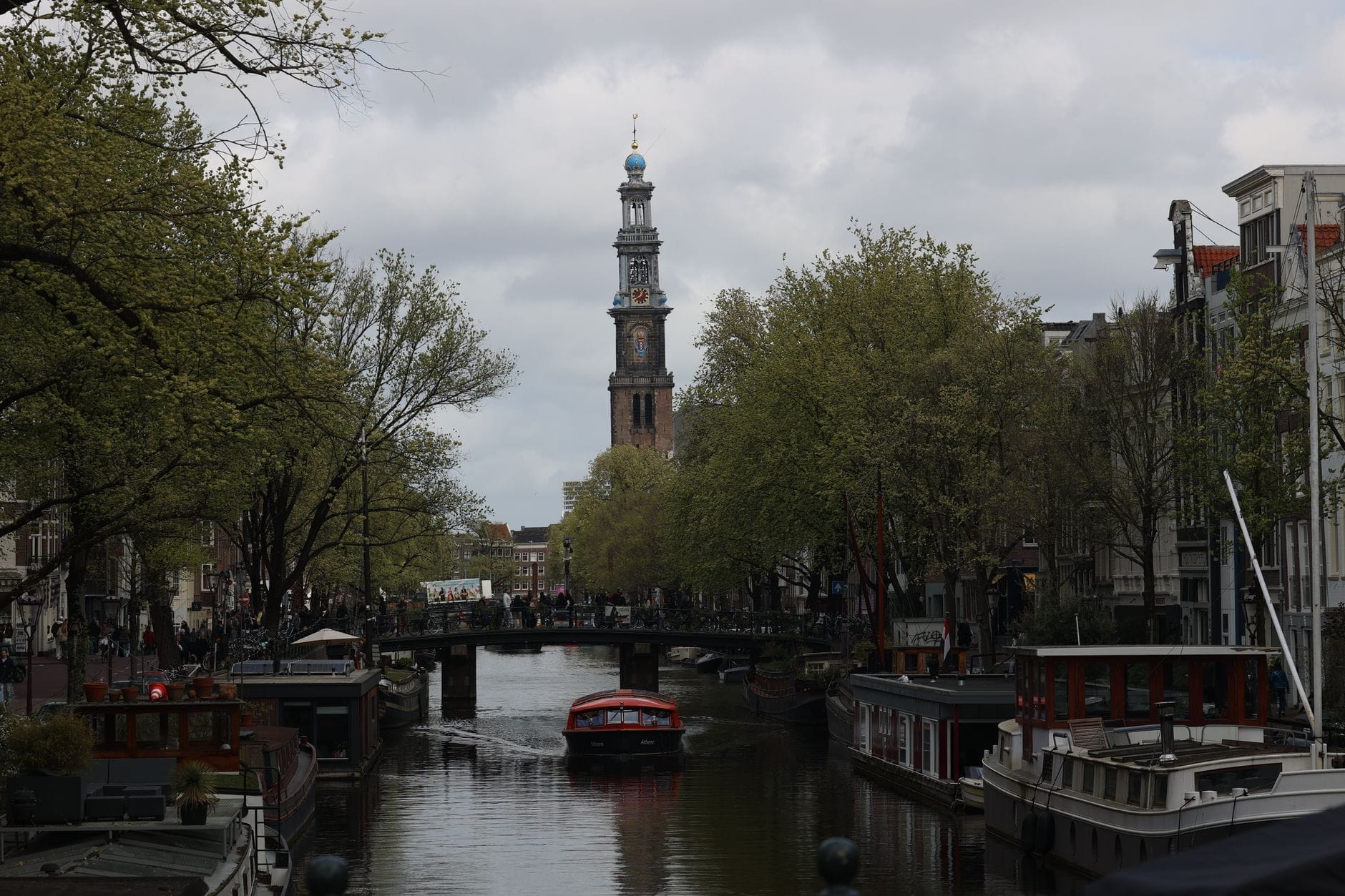 Vista del canal con barcos y la torre de la Westerkerk en Ámsterdam, flanqueada por árboles y edificios tradicionales en un día nublado.