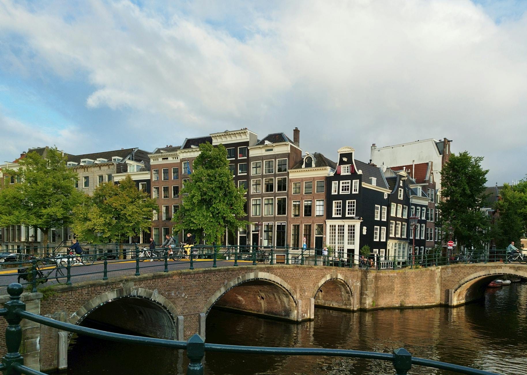Vista clásica de un canal de Ámsterdam con puente arqueado, hilera de casas estrechas y árboles en un día soleado, perfecta para explorar la ciudad con poco presupuesto.