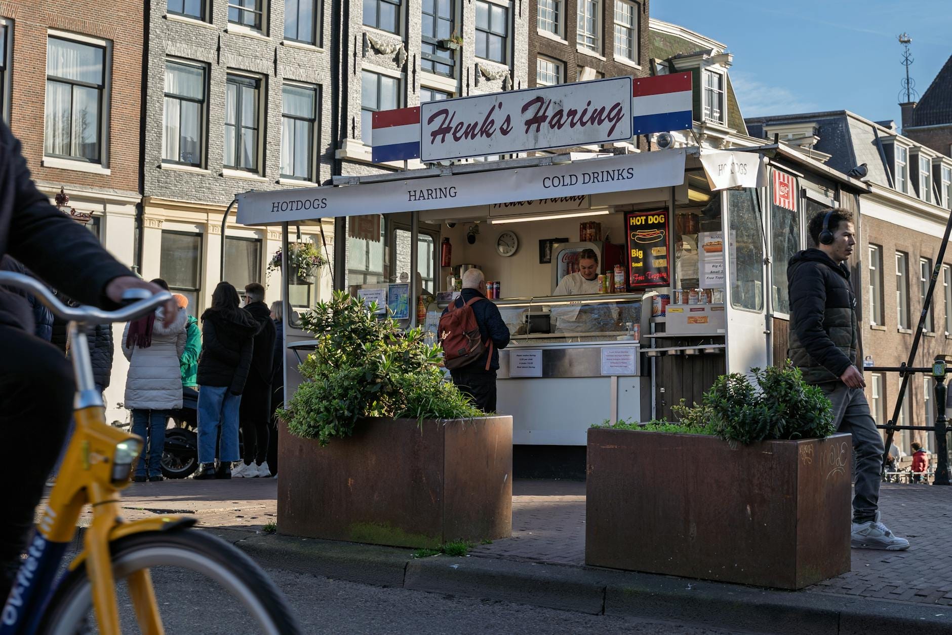 Puesto de comida callejera en Amsterdam con personas pidiendo snacks asequibles cerca de edificios tradicionales.