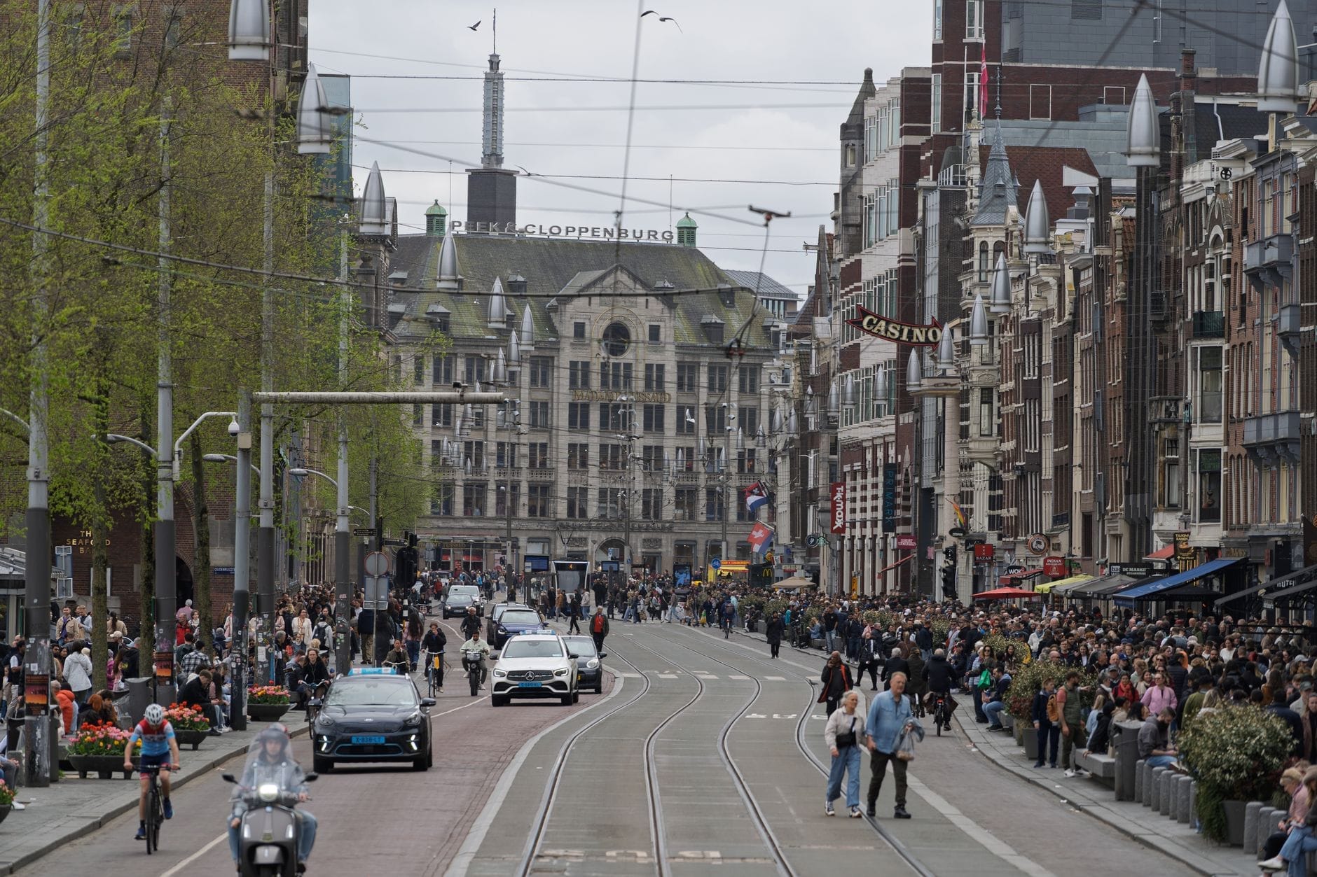 Concurrida calle de Ámsterdam con multitudes, ciclistas, coches y tranvías en un día nublado, mostrando patrones de aglomeración y actividad urbana.