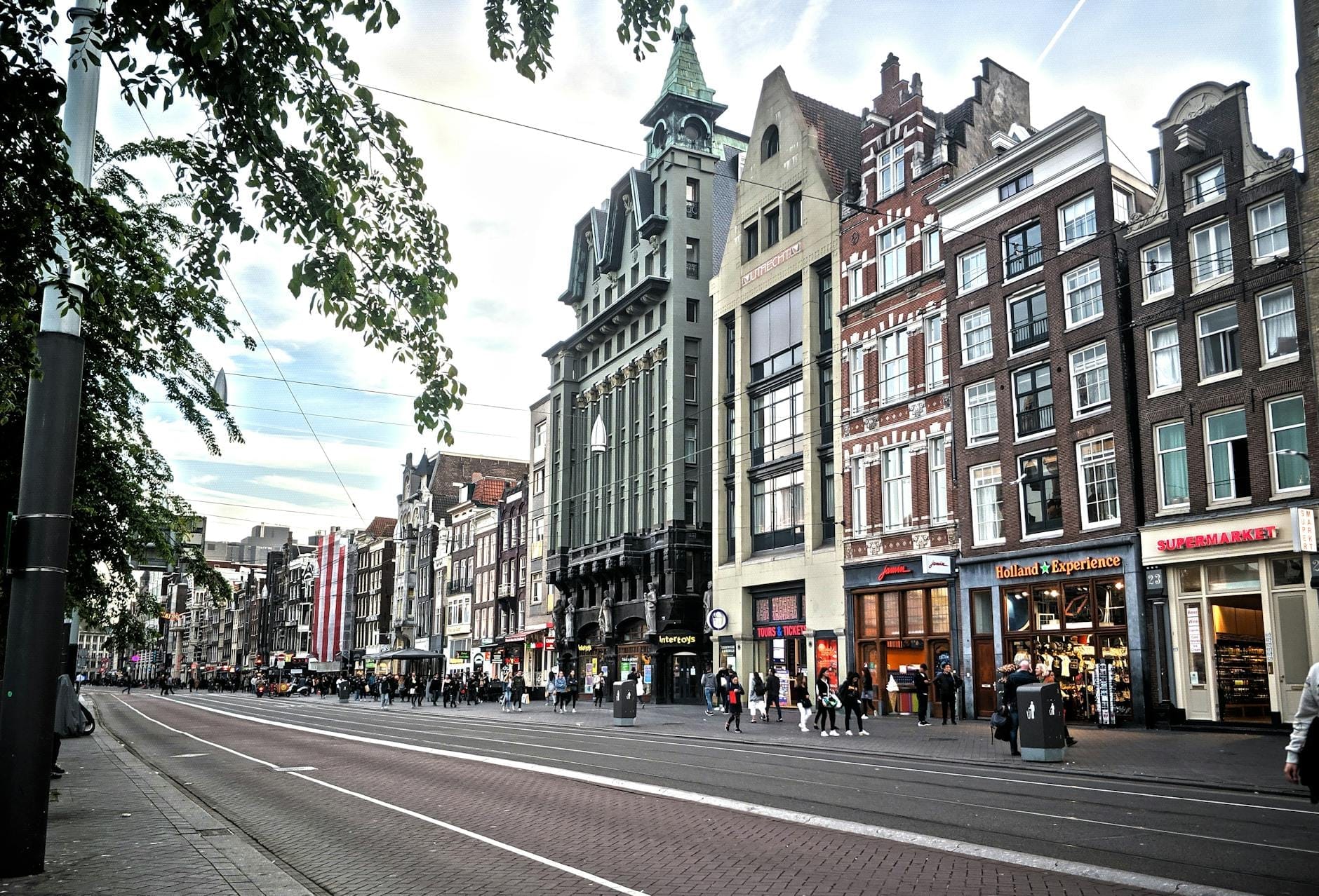 Calle de Amsterdam bordeada de edificios históricos y una variedad de animadas tiendas, con peatones visibles en las aceras y vías de tranvía que pasan por allí.