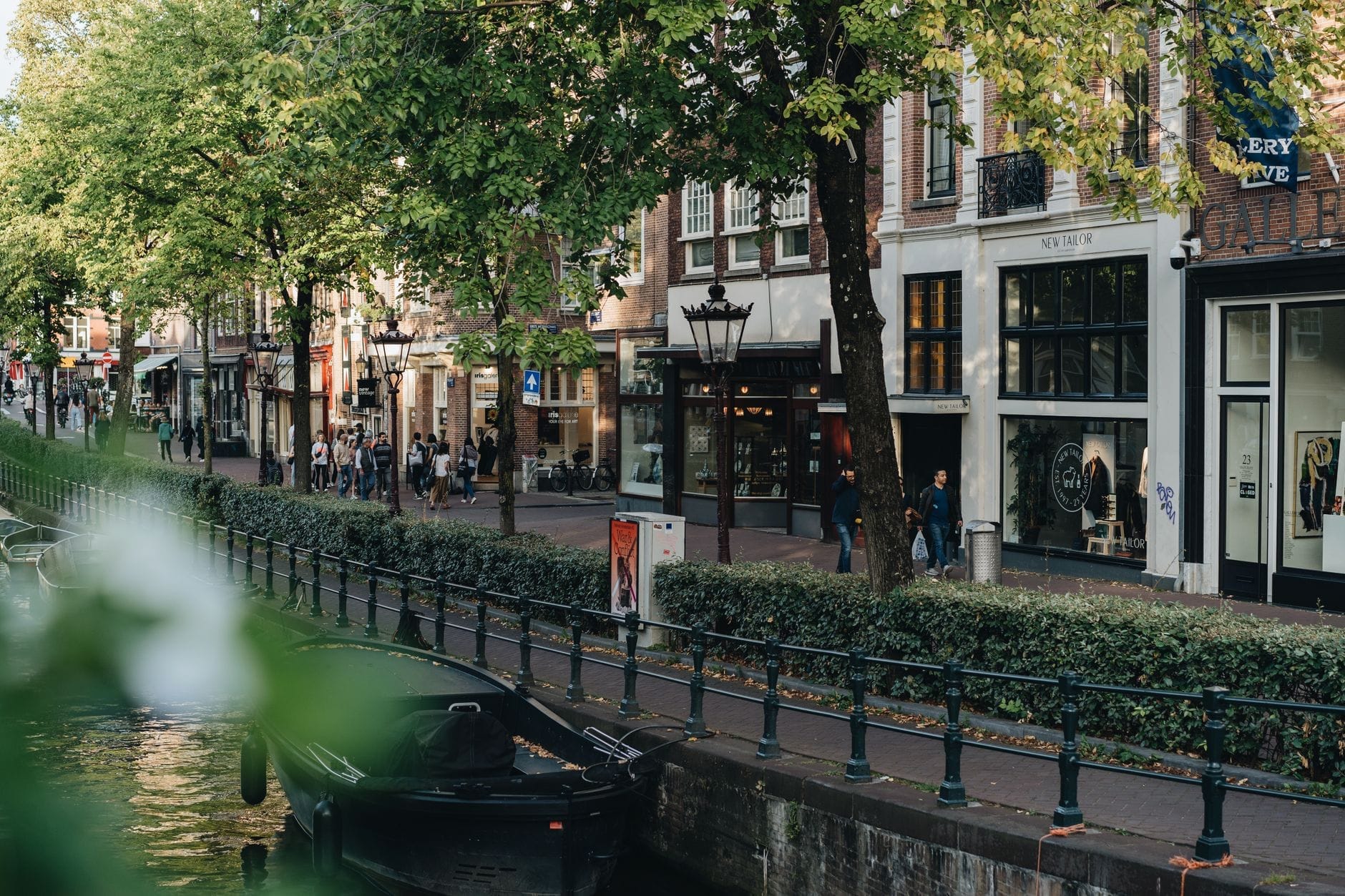 Pintoresca calle comercial de Ámsterdam junto a un canal, con boutiques, árboles y peatones visibles en la acera.