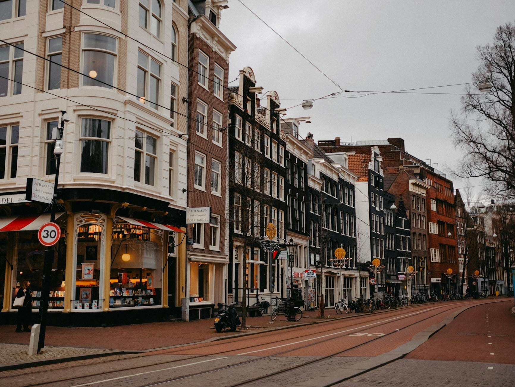 Vista de una calle de Amsterdam con edificios clásicos holandeses, líneas de tranvía y una tienda en la esquina, evocando el ambiente animado de un distrito comercial.