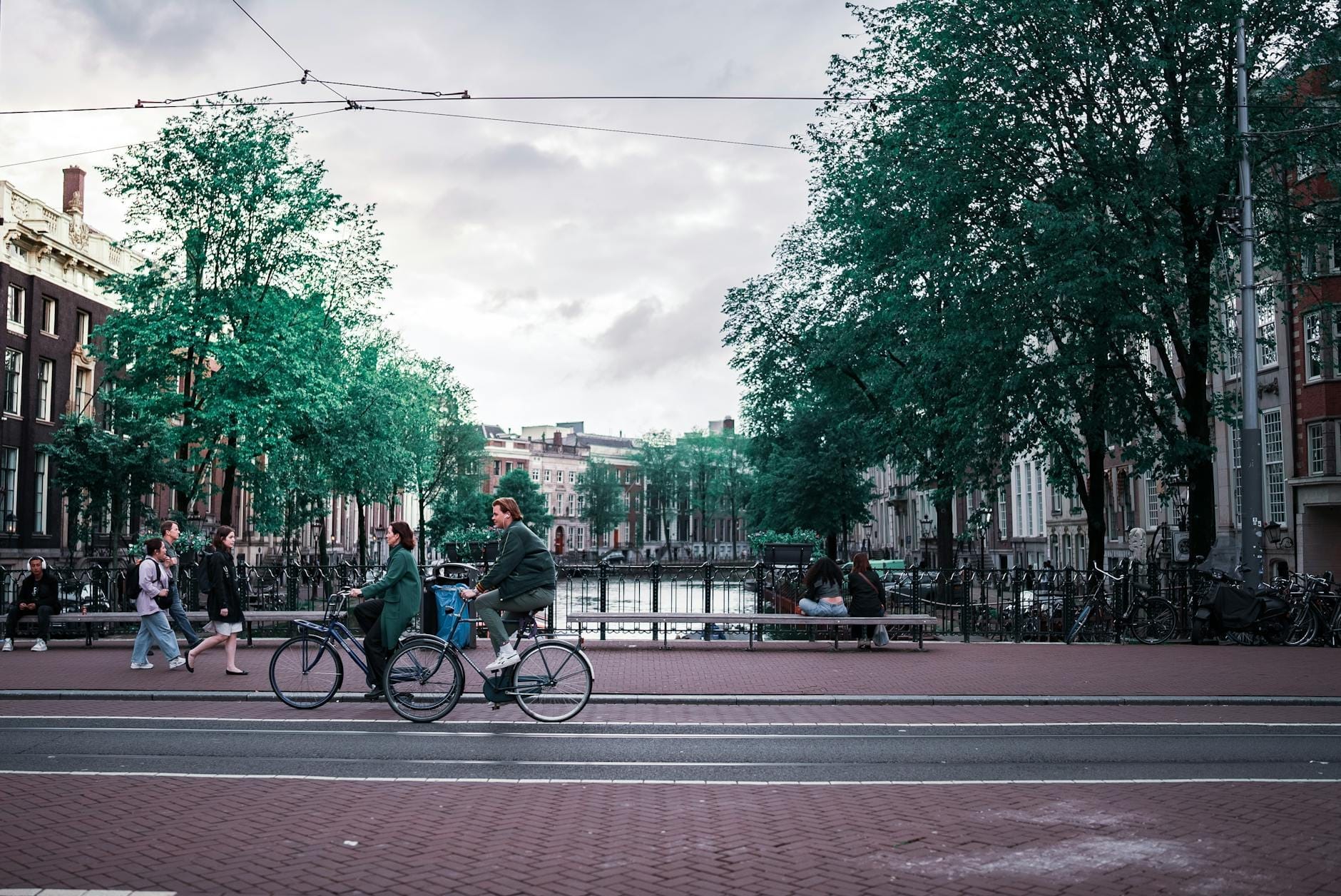 Personas caminando y en bicicleta junto a un canal en Ámsterdam, con casas típicas de canal y árboles al fondo.