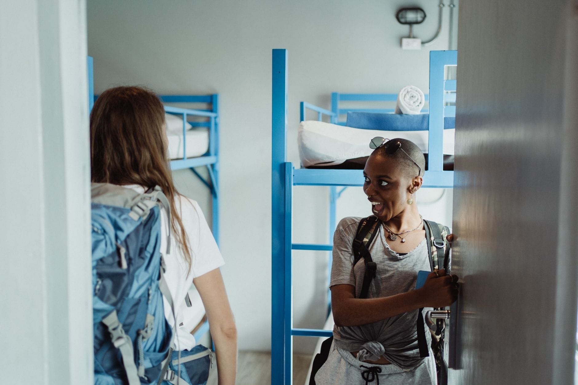 Dos viajeros solitarios con mochilas entran en una habitación de dormitorio de albergue moderno con literas azules, listos para instalarse.