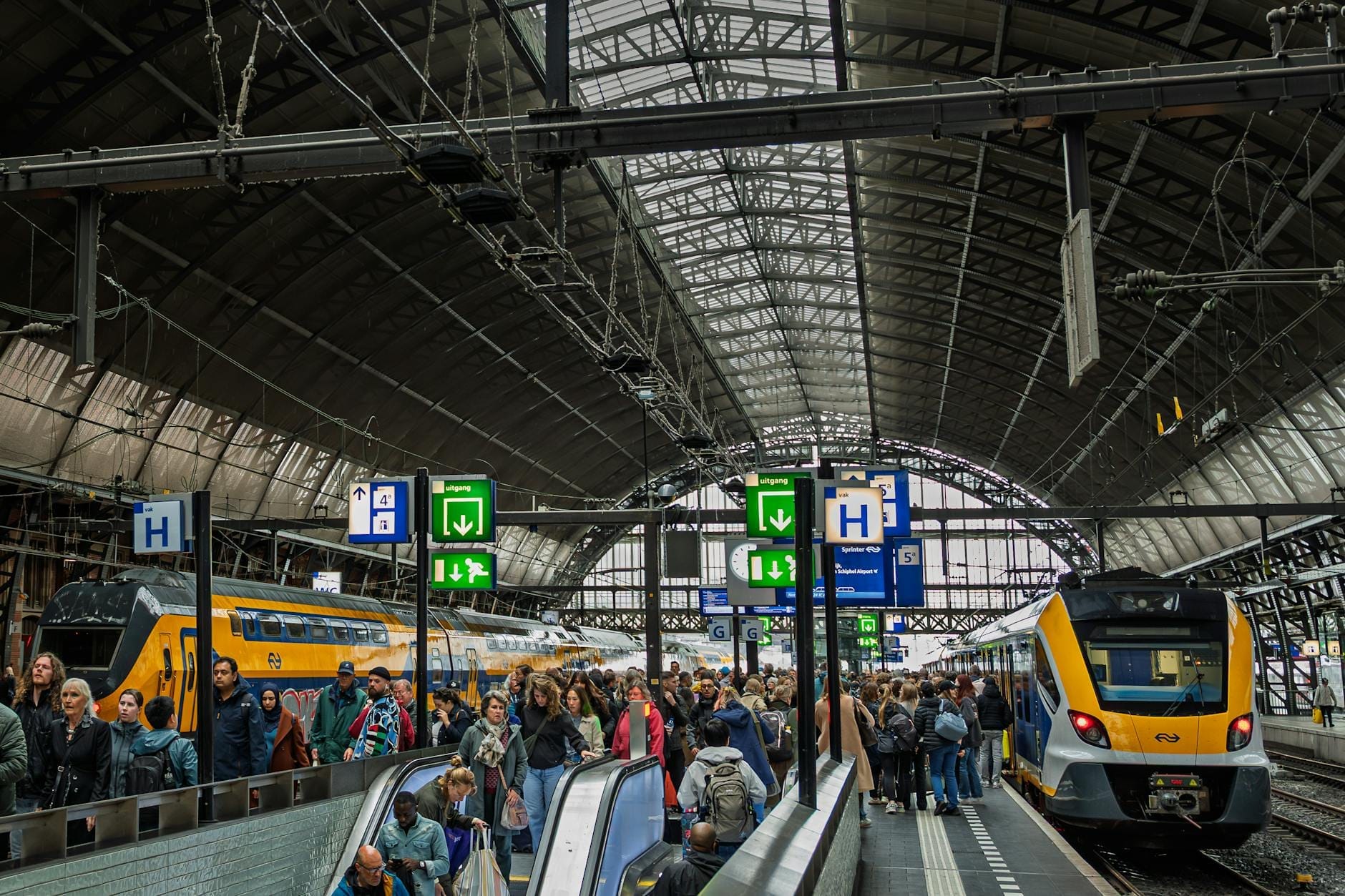 Andén concurrido en la estación de Amsterdam Centraal con dos modernos trenes holandeses bajo la icónica cubierta de cristal abovedada de la estación.