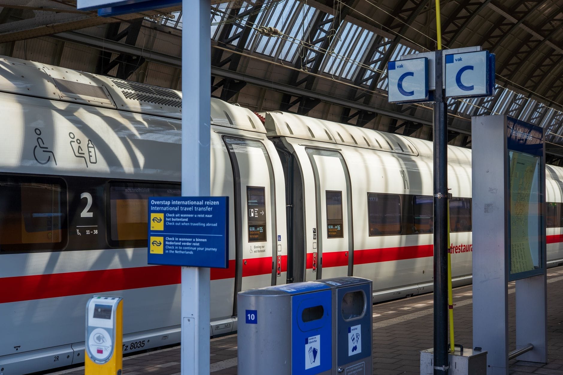 Vista lateral de un moderno tren ICE blanco con franja roja en un andén de la estación Amsterdam Centraal, bajo una gran cubierta de vidrio arqueada.