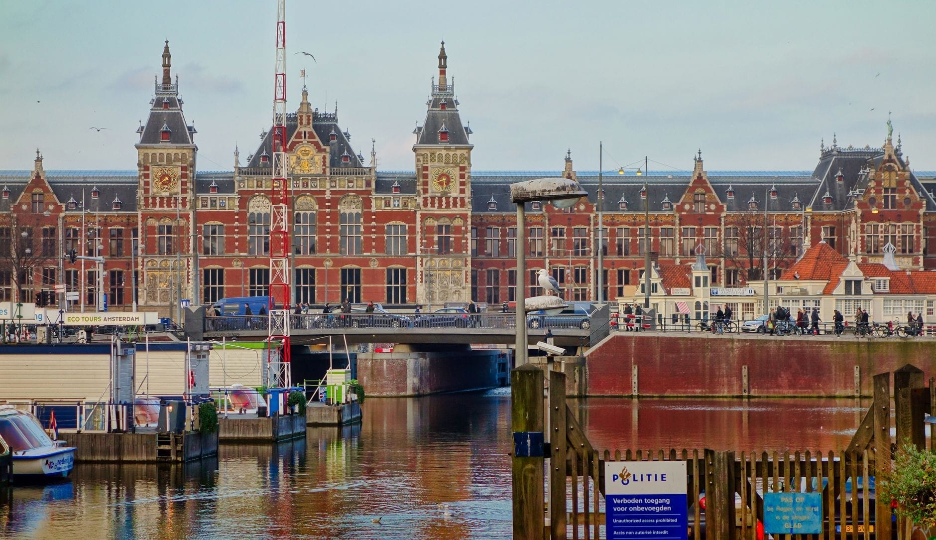 Exterior de la estación Amsterdam Centraal con arquitectura ornamentada, canal de la ciudad en primer plano y personas visibles cerca de la entrada.
