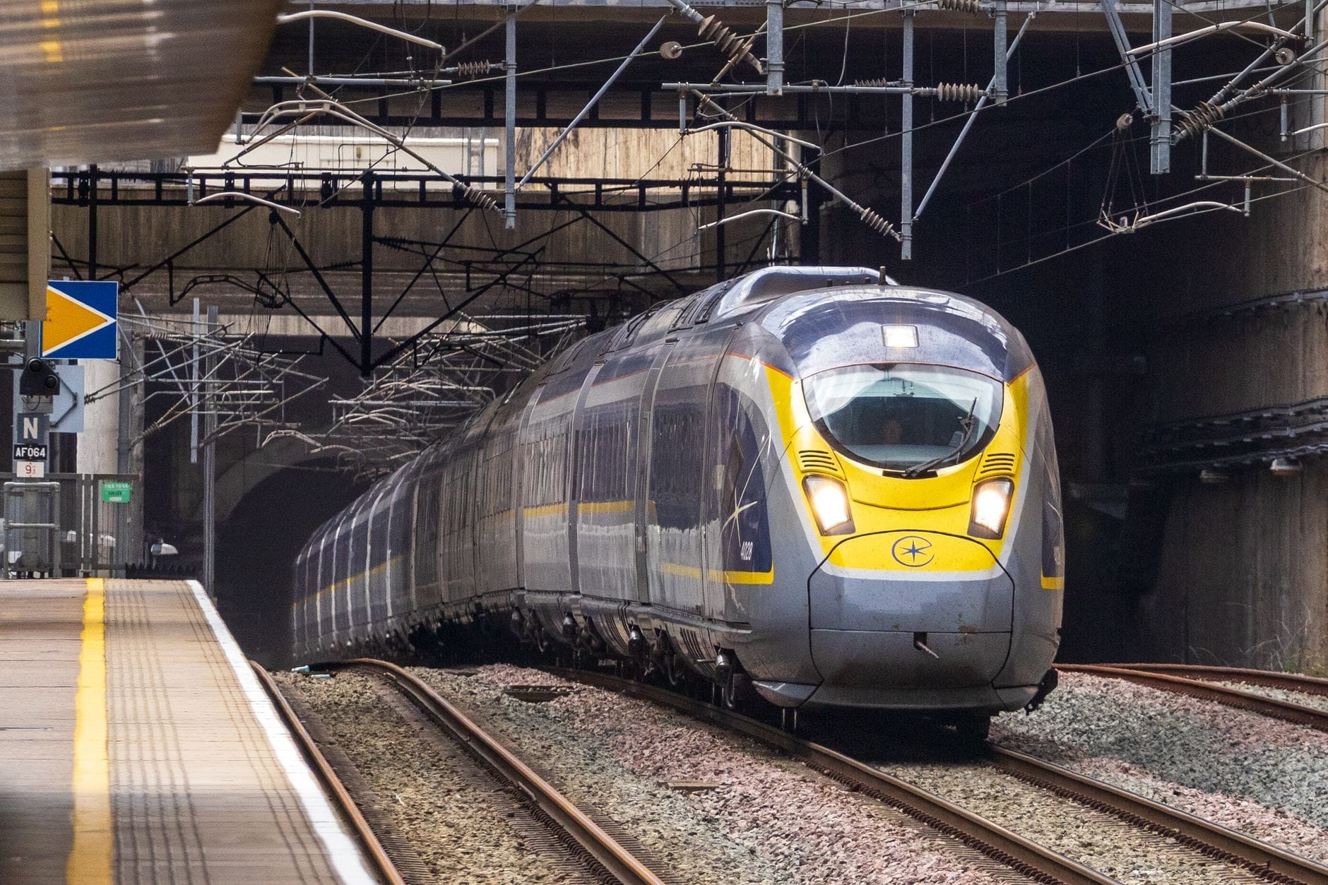 Un moderno tren de alta velocidad Eurostar saliendo de un túnel hacia las vías, con el andén de una estación visible a la izquierda bajo cables aéreos.