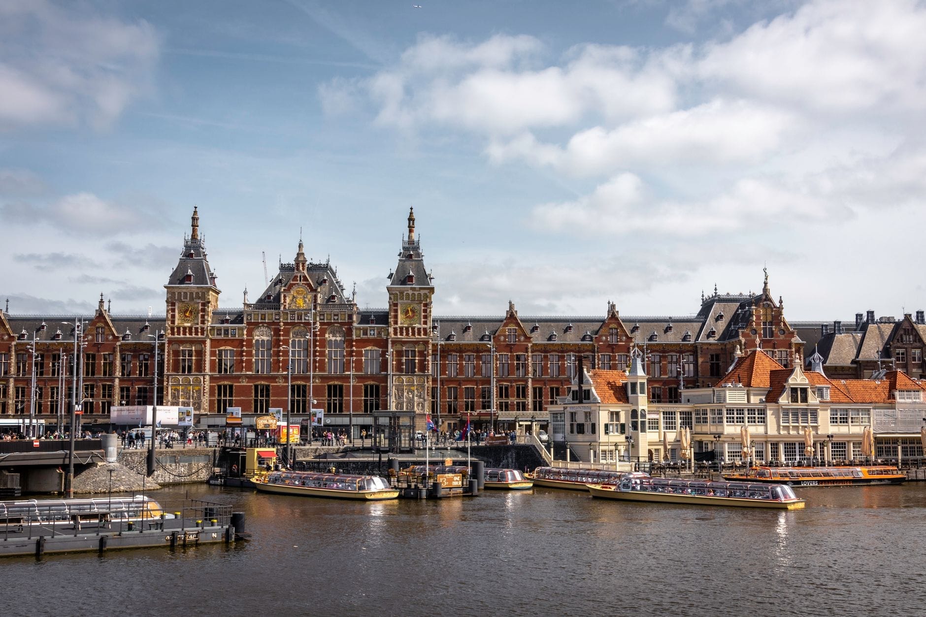 Vista amplia de la estación Amsterdam Centraal con barcos en el canal frente a ella, mostrando la arquitectura de la estación en un día soleado.