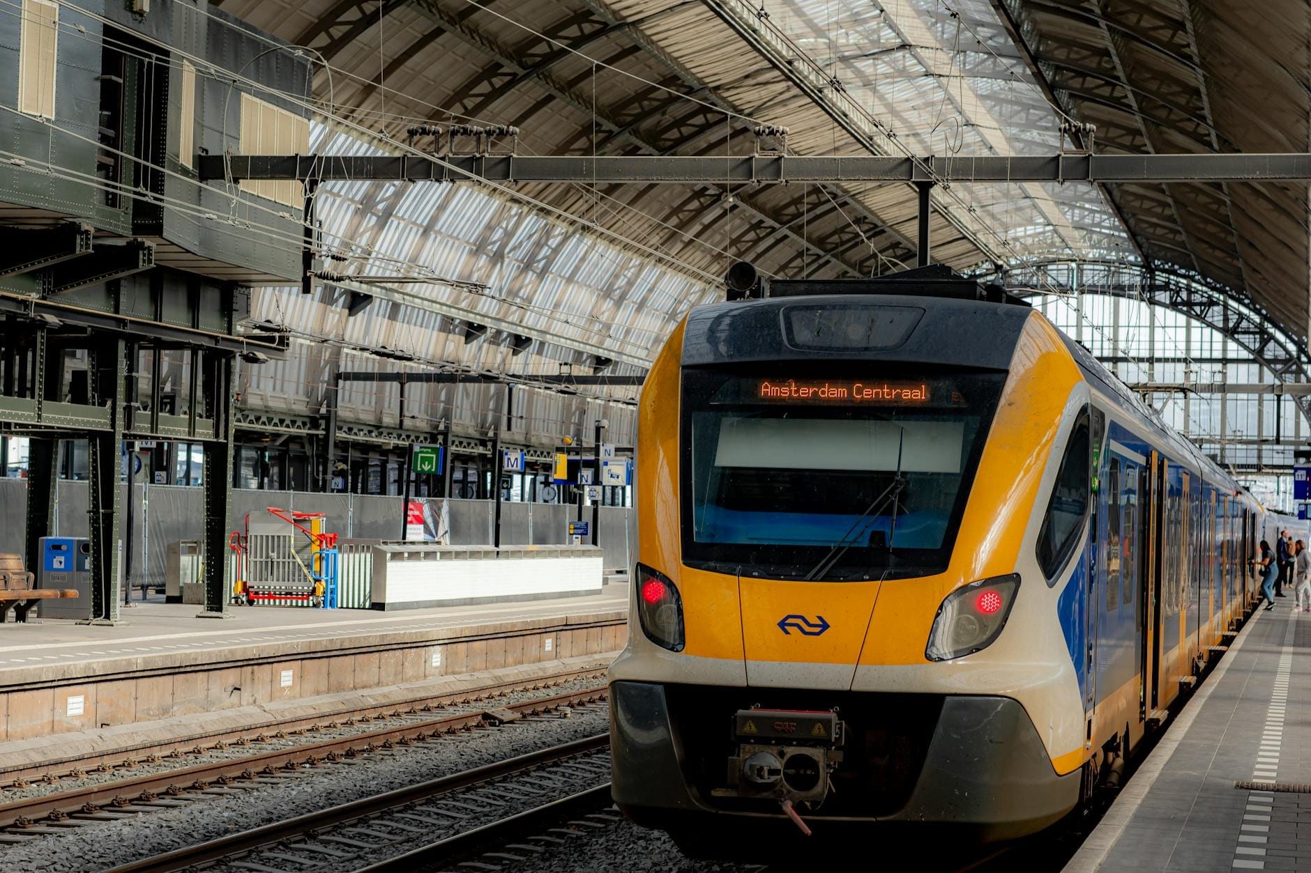 Tren holandés moderno amarillo y azul en un andén dentro de la estación Amsterdam Centraal bajo un techo de cristal arqueado.