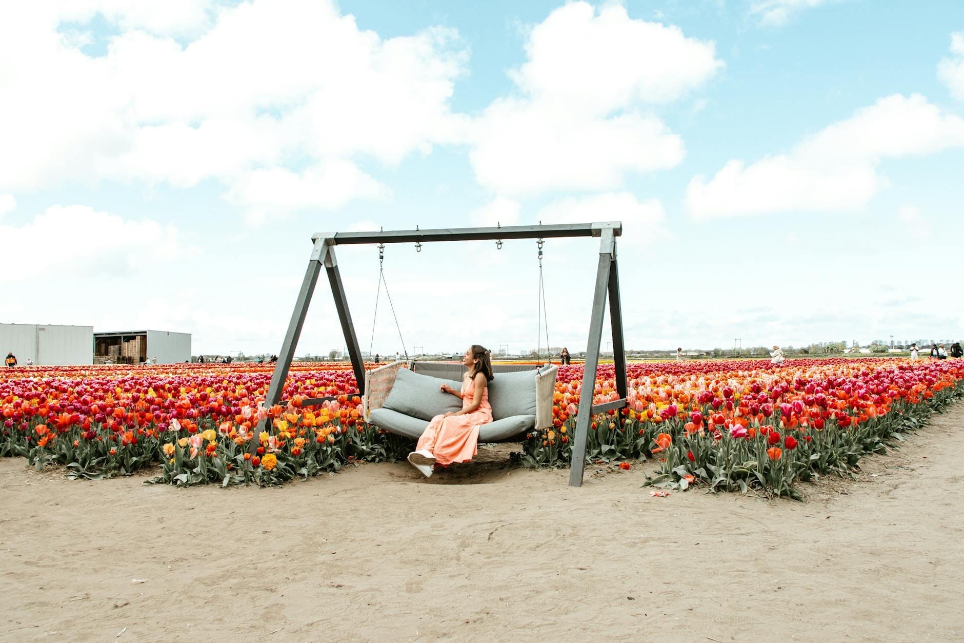 Una mujer con vestido melocotón se sienta en un columpio rodeada de vibrantes campos de tulipanes bajo un cielo azul, evocando la belleza de la temporada de tulipanes en Ámsterdam.