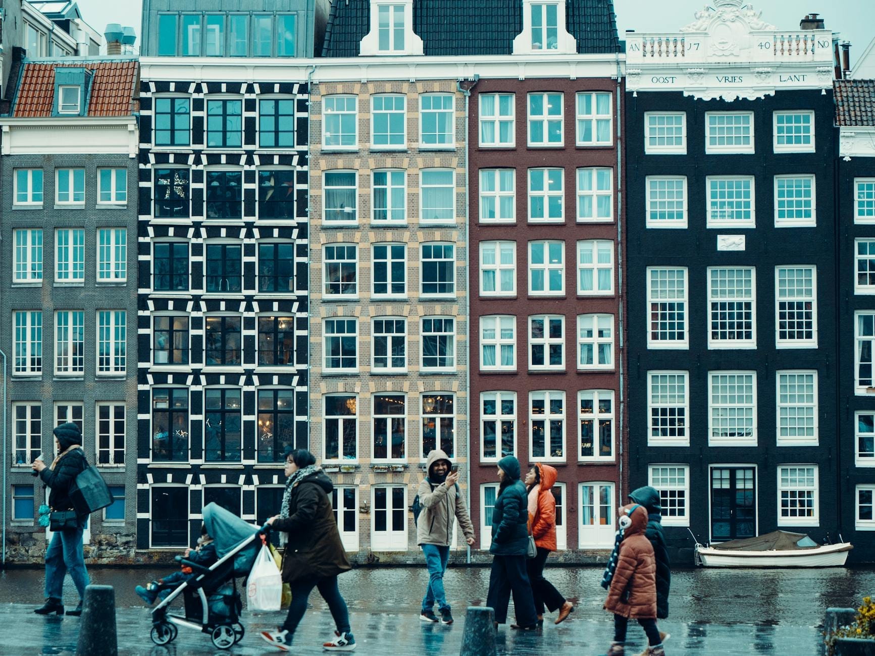 Personas caminando frente a las características casas estrechas de los canales de Ámsterdam, con fachadas de colores y reflejos en el pavimento mojado, una imagen que captura el encanto urbano de la ciudad.