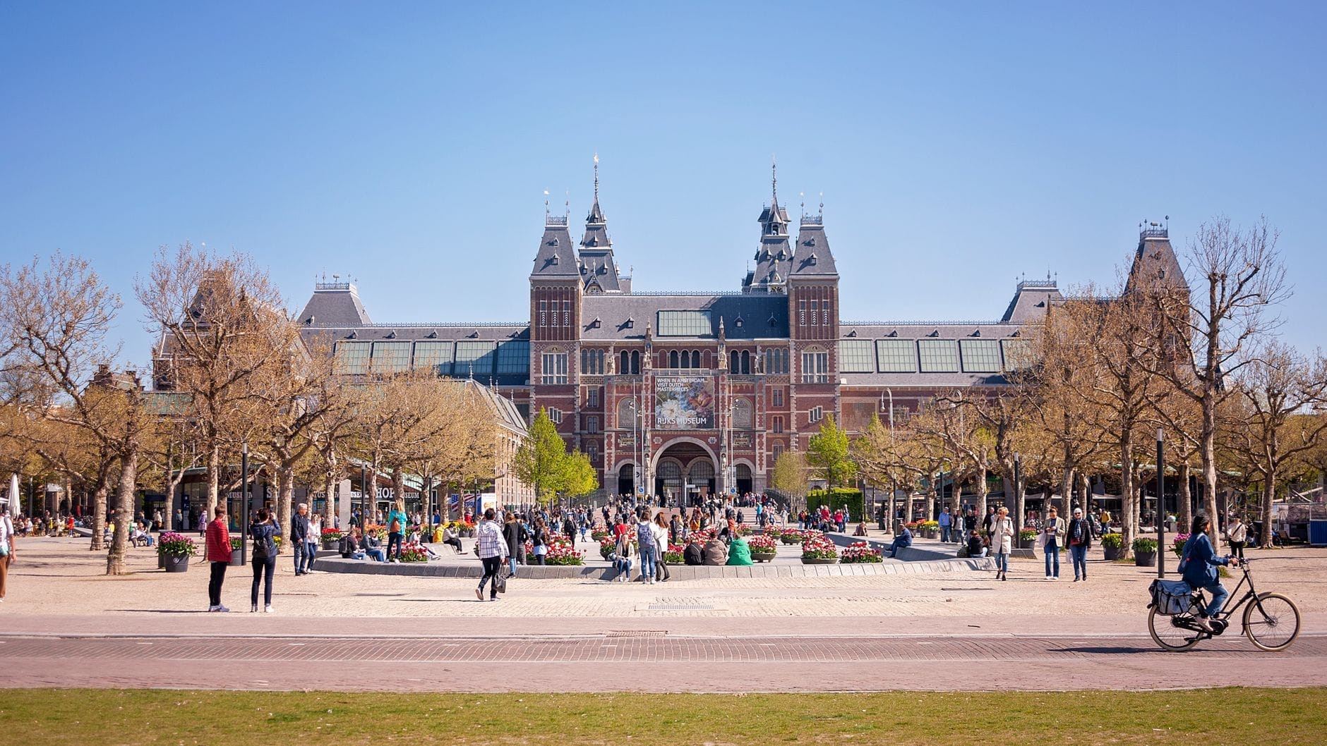 Cảnh nhìn Rijksmuseum từ Museumplein với người đi bộ và đạp xe ở phía trước vào một ngày nắng đẹp.