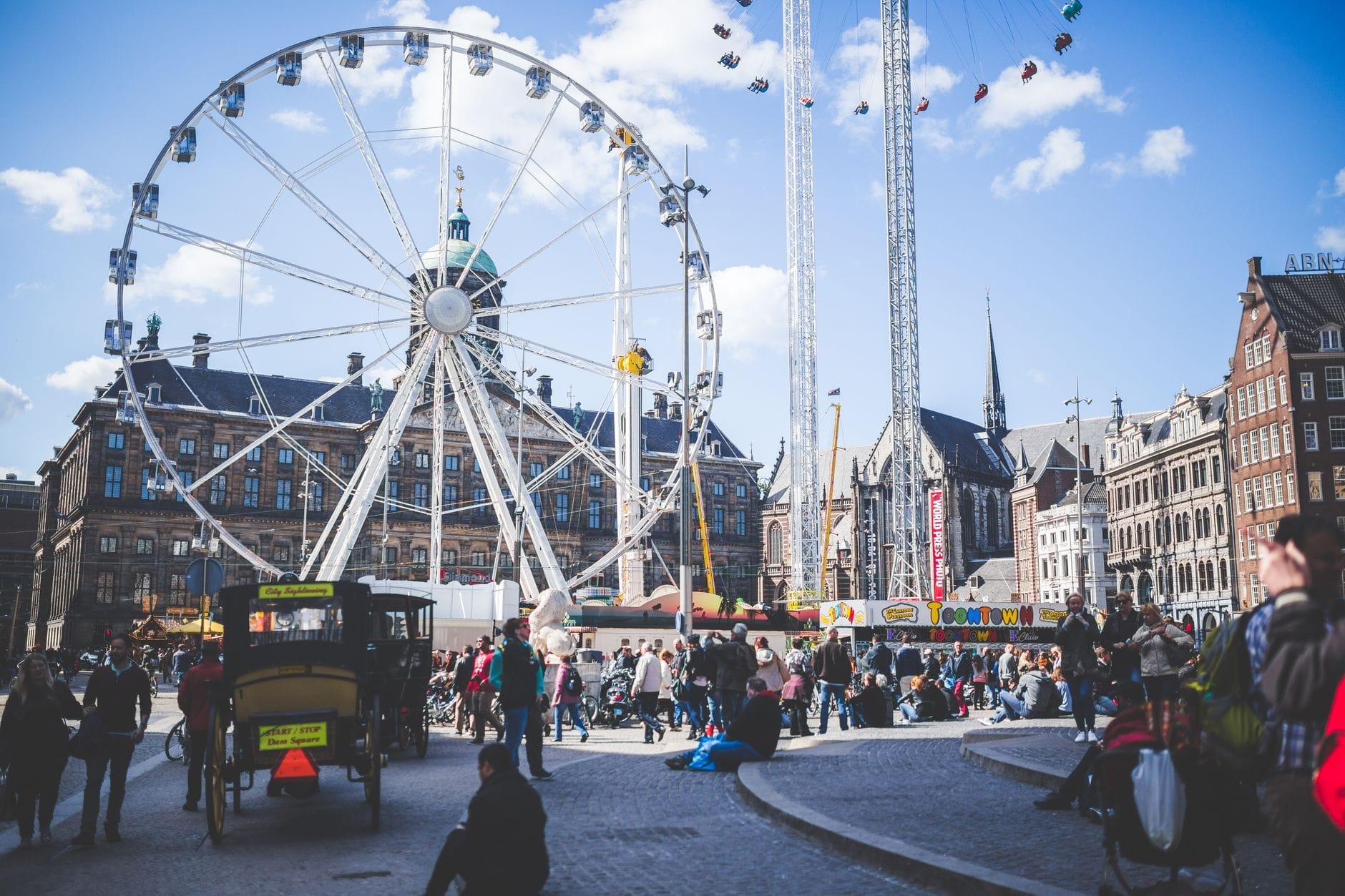 Animada plaza de Ámsterdam con una gran noria, edificios históricos y numerosas familias disfrutando del día bajo un cielo azul brillante.