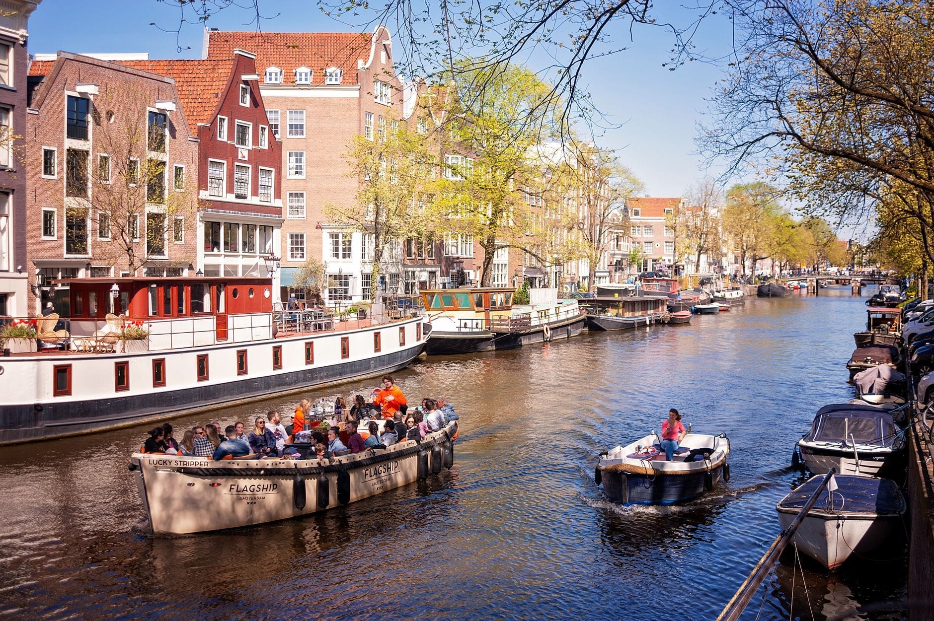 Barco turístico abierto con personas disfrutando de un paseo por un canal de Amsterdam, rodeado de casas históricas y otros barcos en un día soleado.