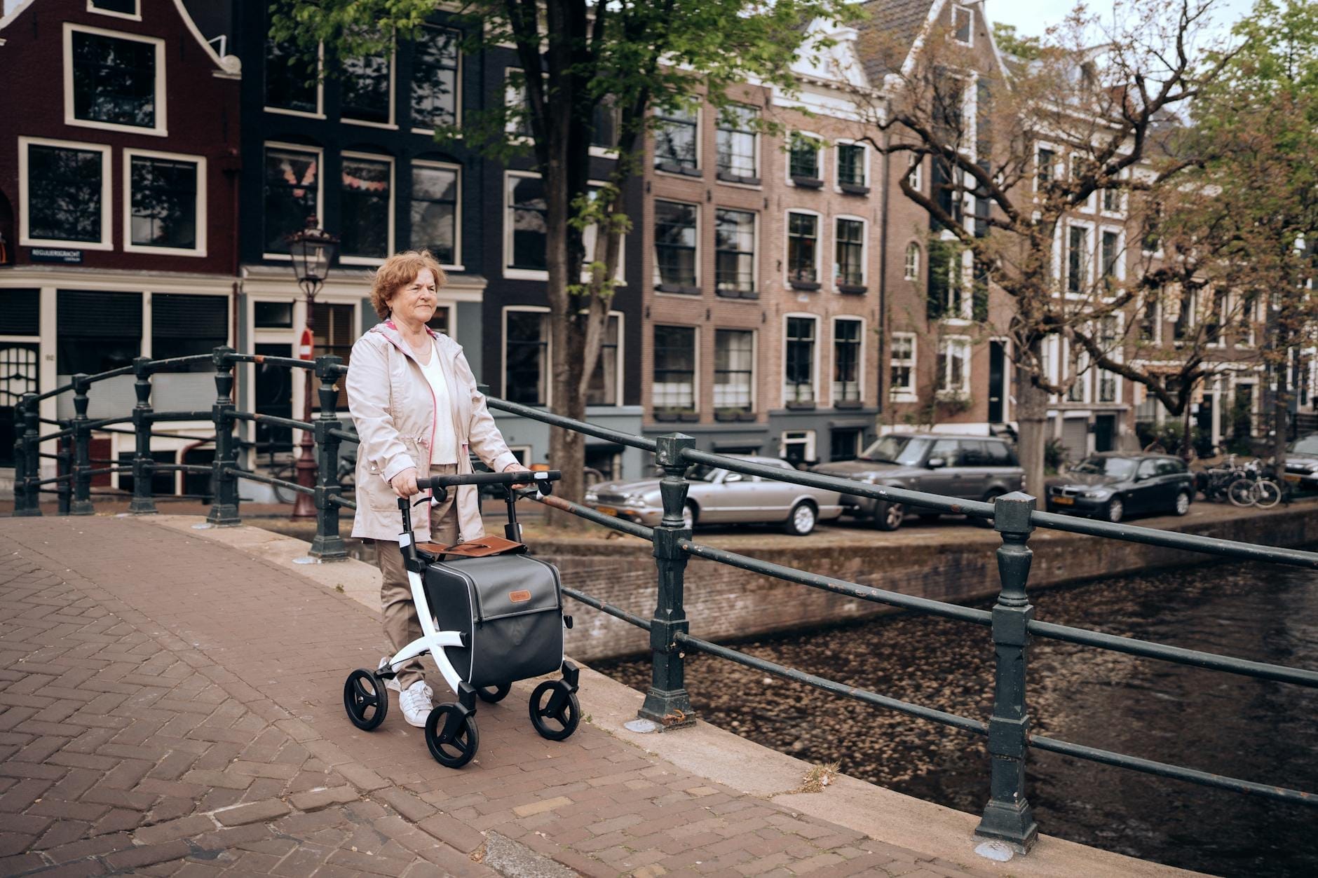 Mujer con andador cruzando un puente sobre un canal de Ámsterdam, con típicas casas de canal y bicicletas al fondo.