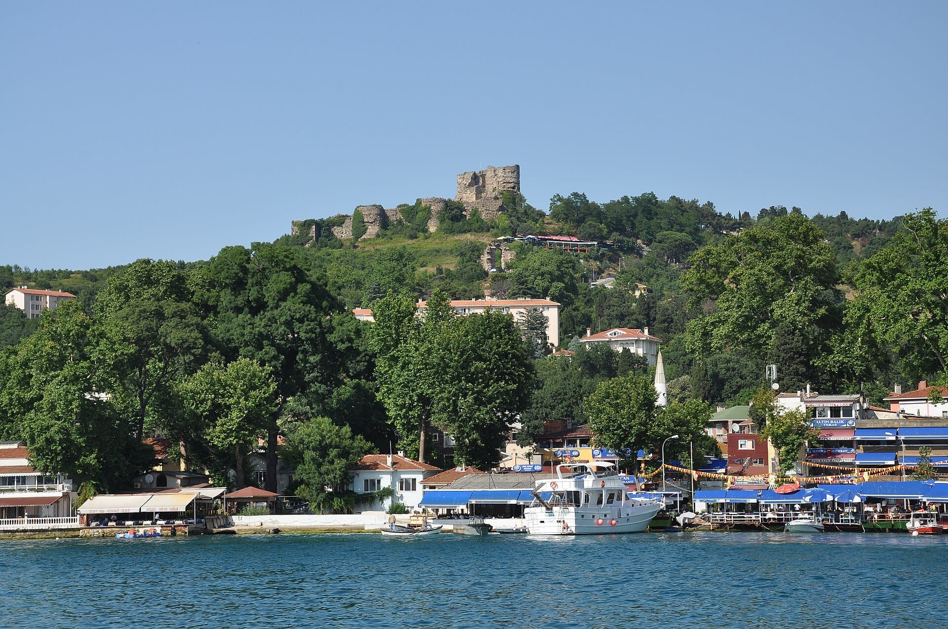 Vista del Castello di Yoros in cima a una collina verde che domina il colorato villaggio e i pescherecci di Anadolu Kavağı lungo le rive del Bosforo.