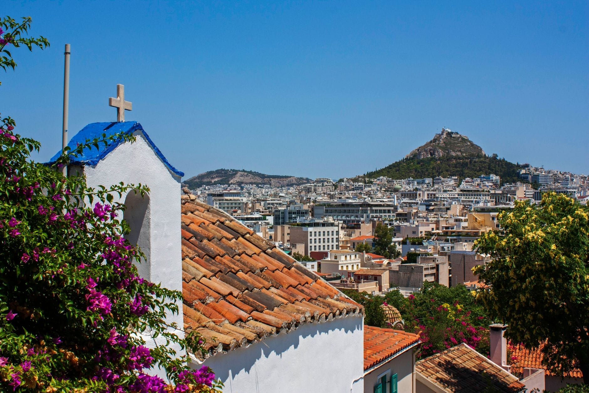 Veduta soleggiata di case imbiancate a calce con tetti in terracotta, una cappella dalla cupola azzurra, bouganvillee in fiore e il panorama di Atene con una collina sullo sfondo.
