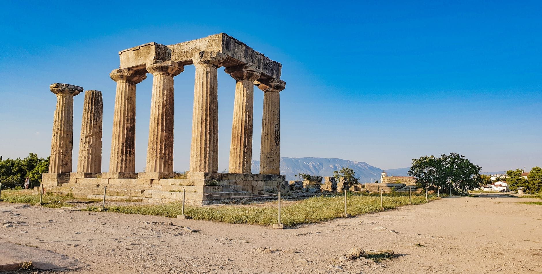 Vista grandangolare delle rovine del tempio greco antico di Corinto, con sette colonne doriche ancora in piedi sotto un cielo azzurro e colline sullo sfondo.