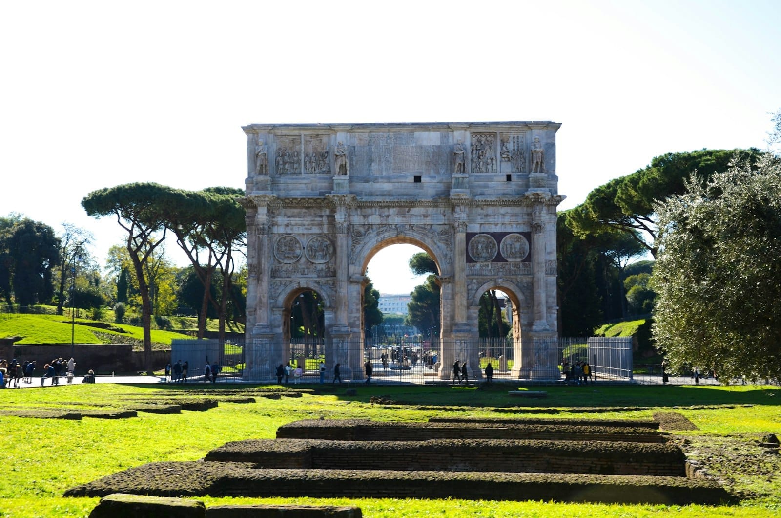 The Arch of Constantine in Rome, a grand ancient Roman triumphal arch set amid bright green grass and framed by stone ruins under a sunny sky.