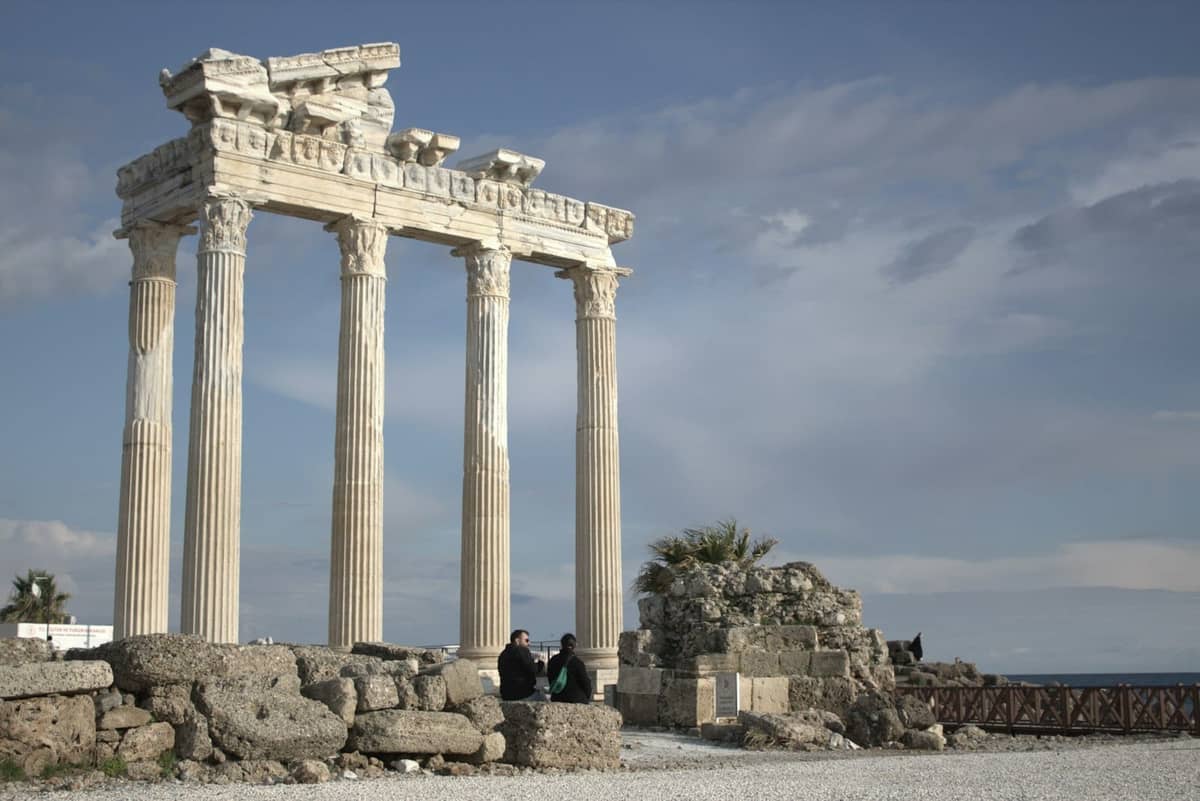 Ancient Roman columns of the Temple of Apollo by the sea in Side, with three people sitting on stone ruins under a cloudy sky.