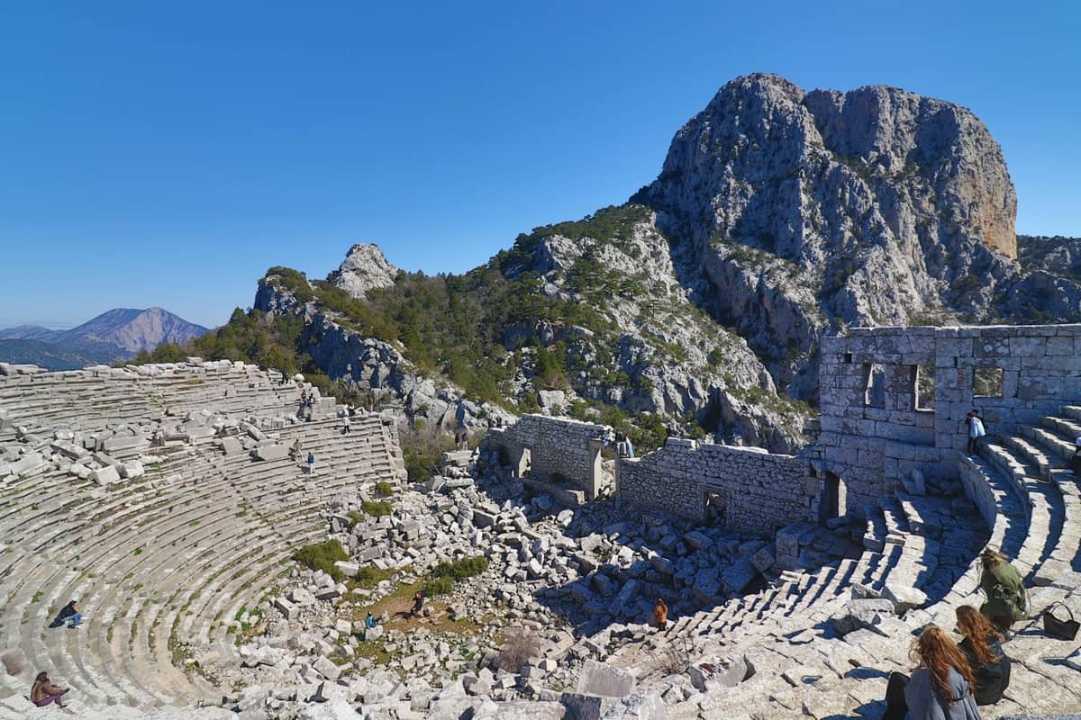 Ancient stone theater ruins at Termessos with dramatic mountain backdrop and clear blue sky, people sitting on steps.