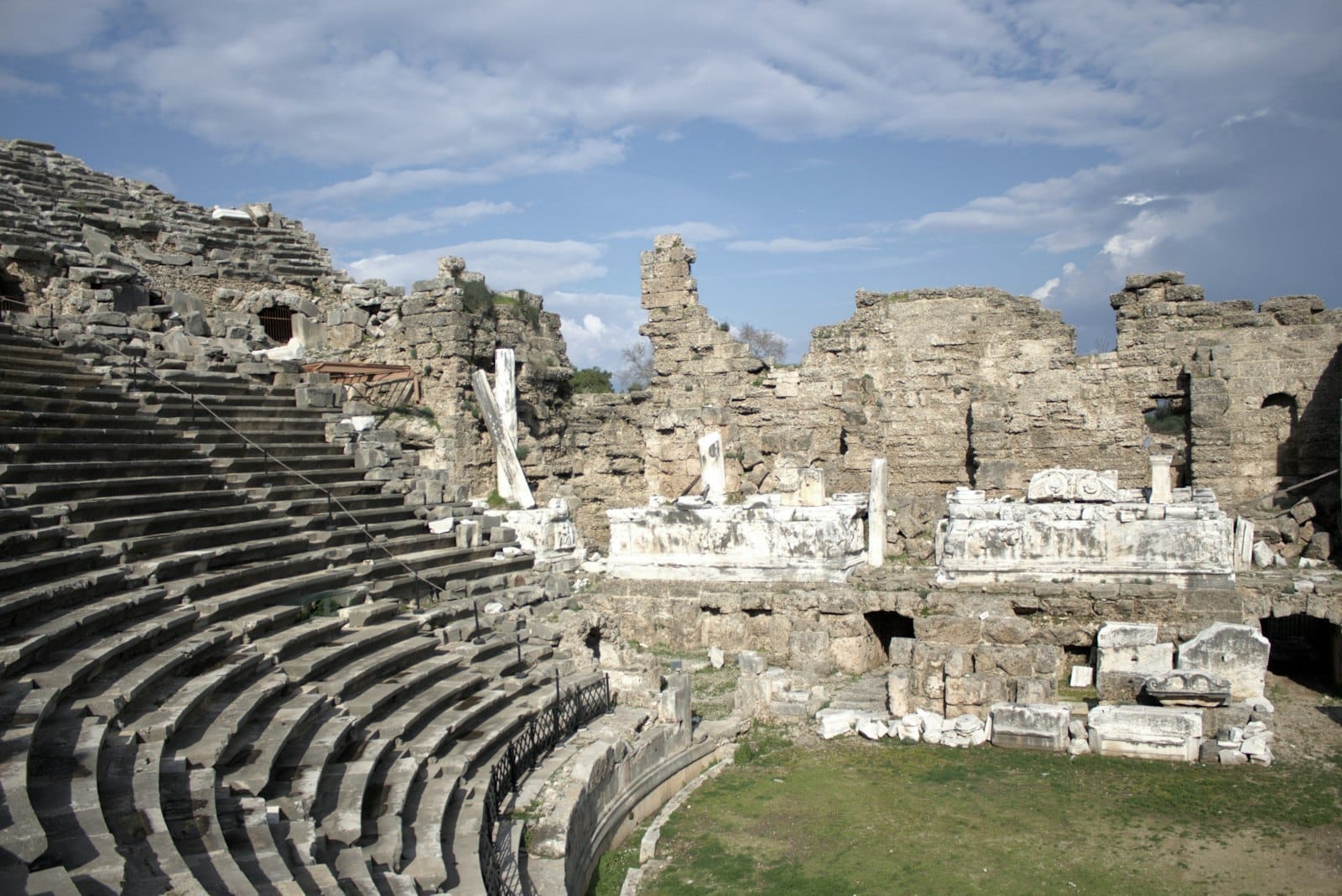 Vista ampla de um teatro romano antigo com arquibancadas de pedra e ruínas sob um céu parcialmente nublado, capturando a grandiosidade dos sítios históricos de Antália.