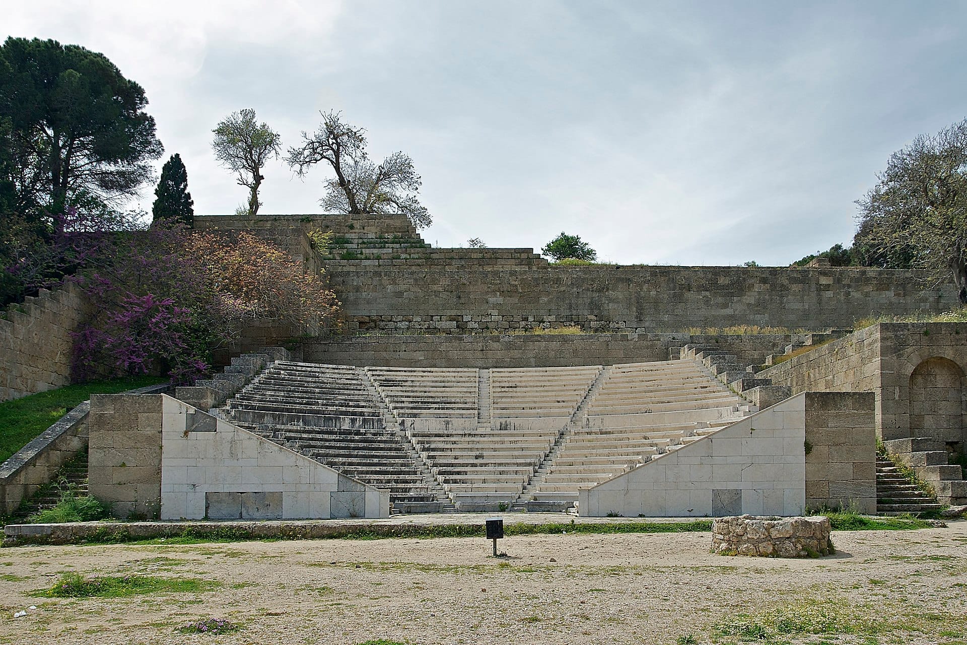 ที่นั่งหินที่ได้รับการบูรณะของสนามกีฬาโบราณแห่ง Rhodes พร้อมขั้นบันไดที่สึกกร่อน ล้อมรอบด้วยกำแพงหินเก่าและต้นไม้กระจัดกระจายใต้ท้องฟ้าที่มีเมฆครึ้ม