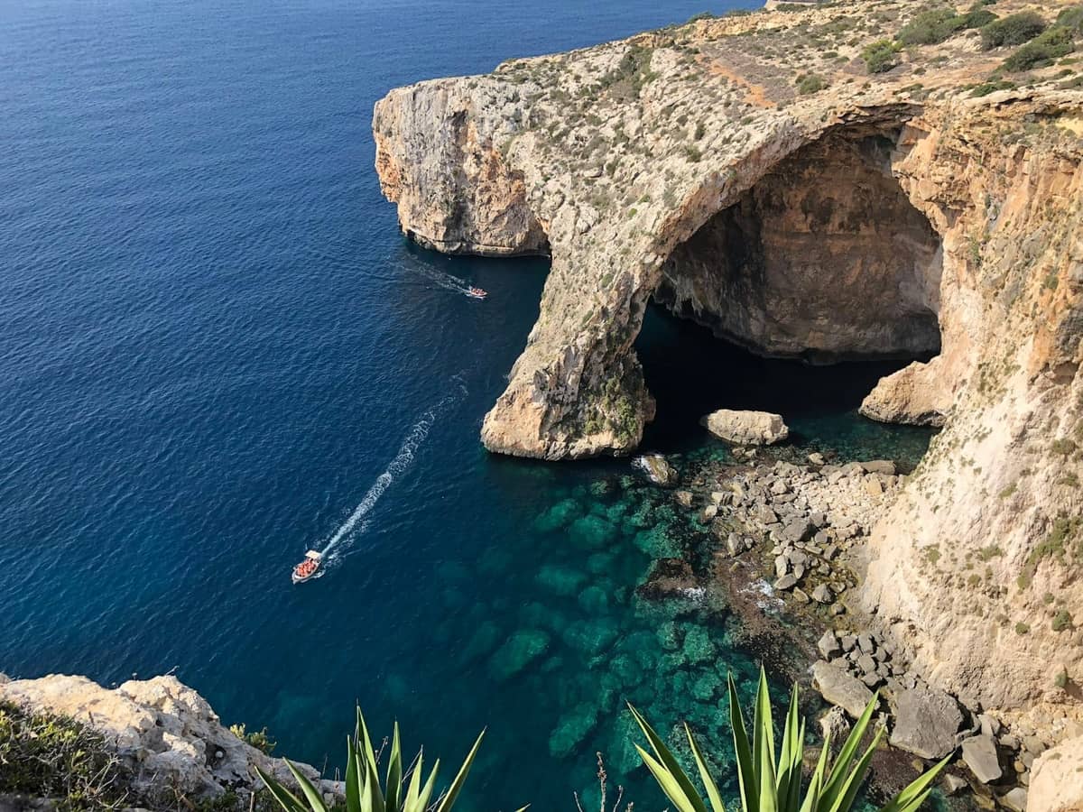 Luftaufnahme der Klippen und des Bogens der Blue Grotto auf Malta mit leuchtend blauem Wasser, Booten und Küstenvegetation.